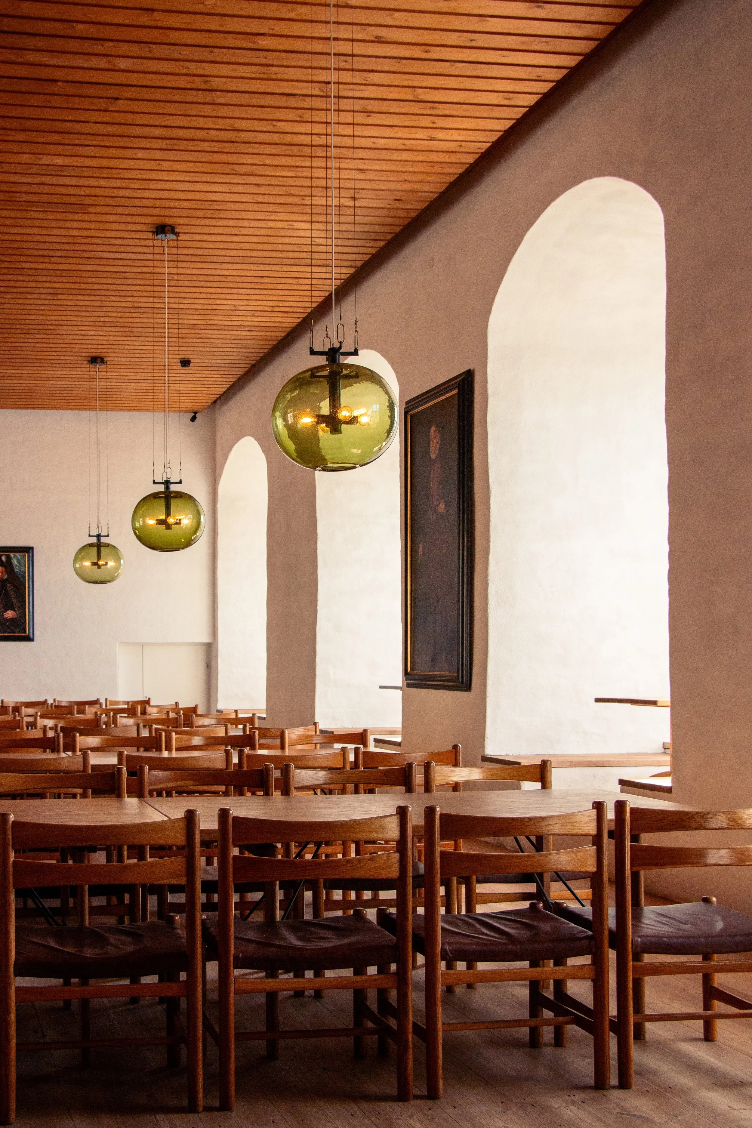 Empty dining hall with wooden tables and chairs, white and gray arched walls, framed paintings, and spherical green pendant lights hanging from a wooden ceiling.