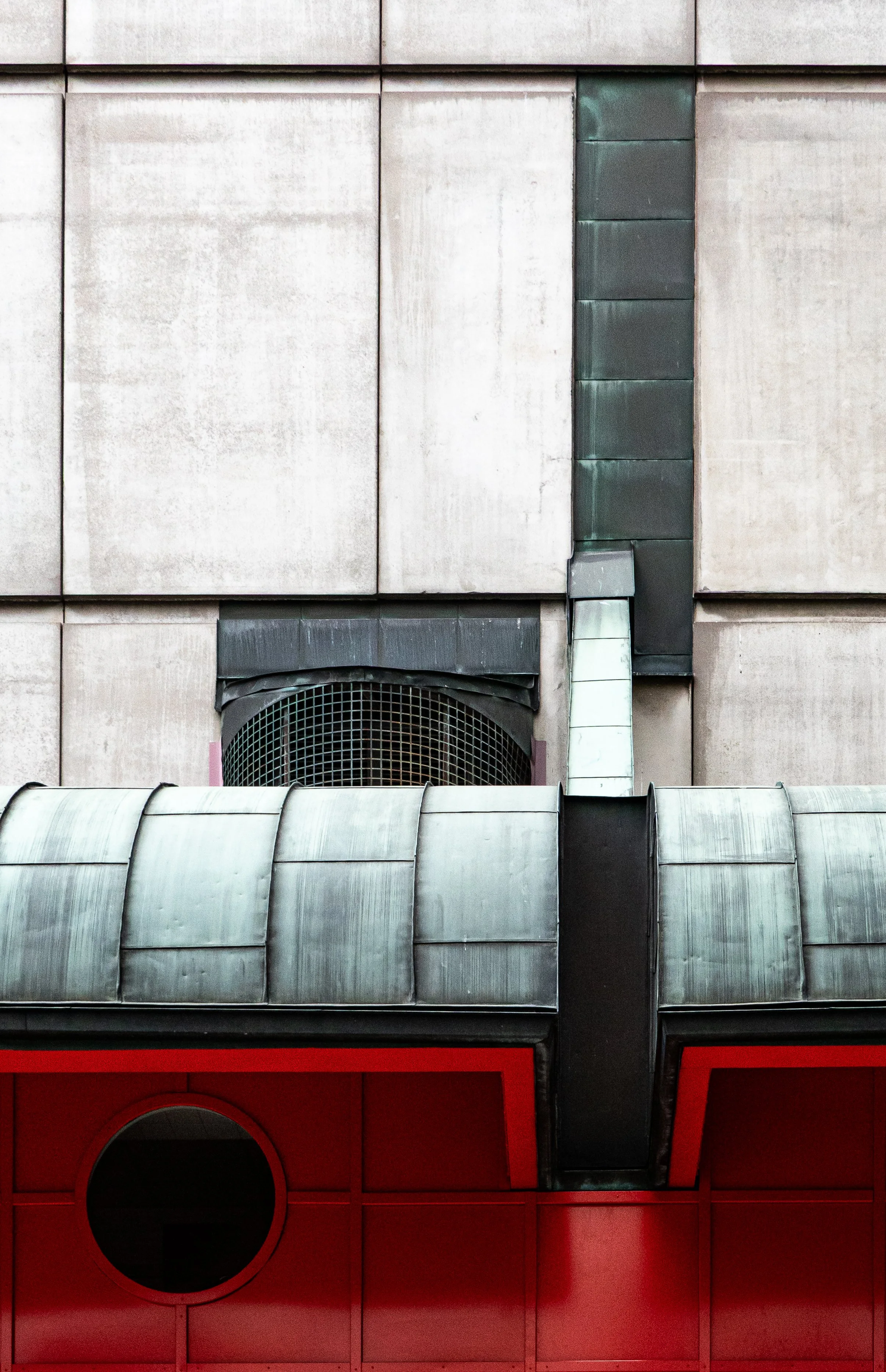 Close-up of a building's exterior with gray concrete panels, a circular vent with a grill, metal cladding, and a red panel with a round window.