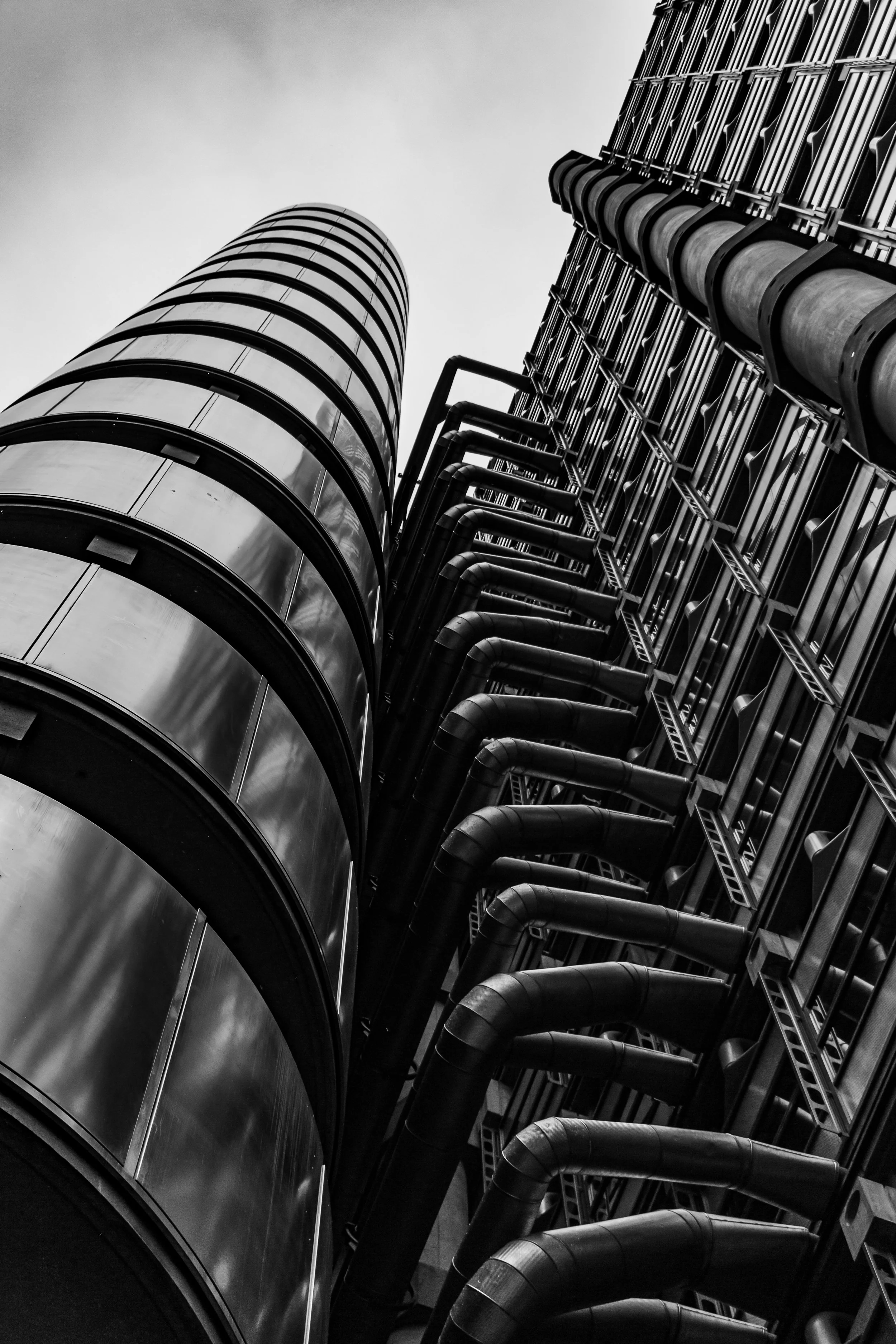 Black and white photo of a modern building with curved glass windows and visible industrial piping on the exterior, taken from a low angle looking up.