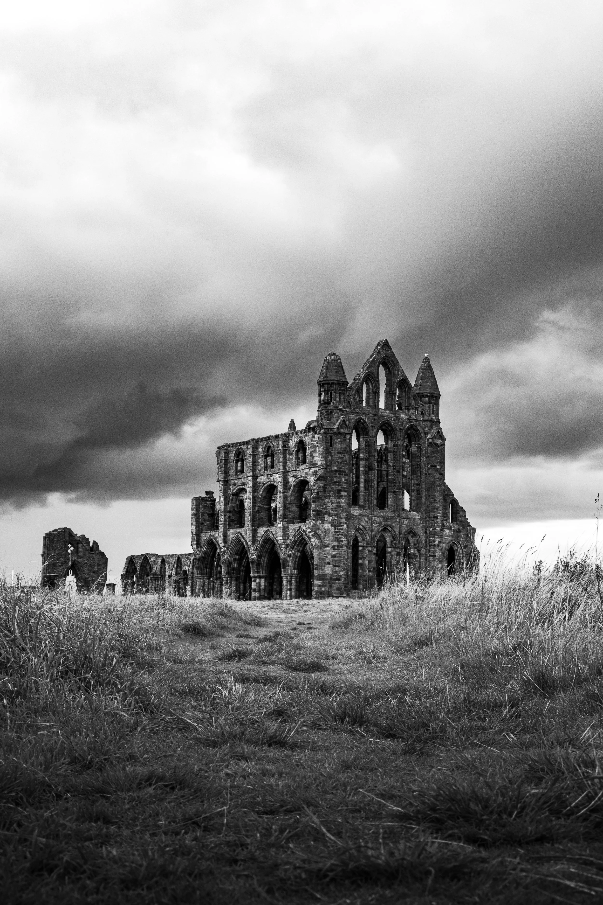 Black and white photo of an abandoned, ruined Gothic church with tall, pointed windows, surrounded by overgrown grass under a cloudy sky.