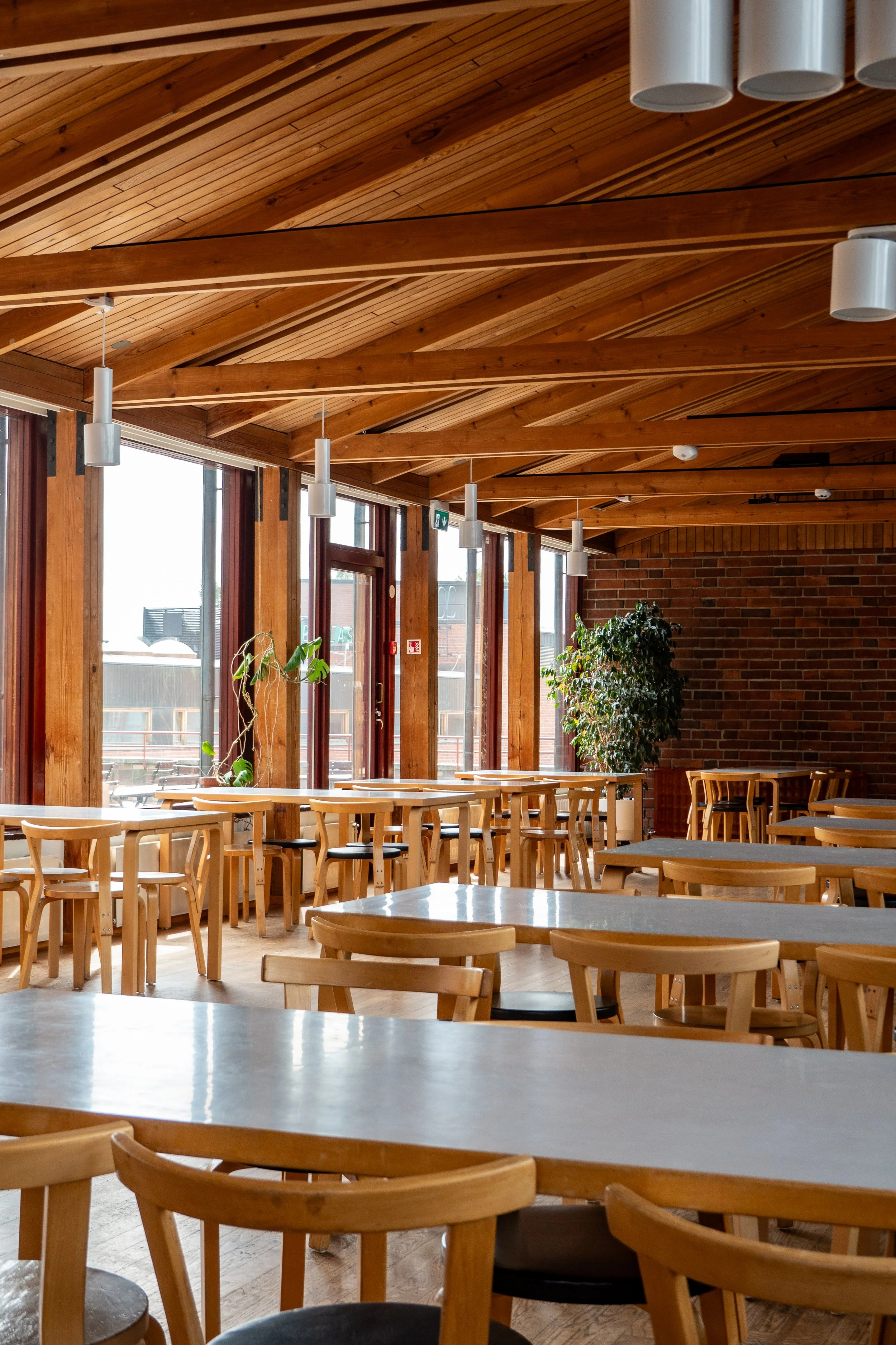 Interior of a modern cafe with wooden tables and chairs, large windows, wooden ceiling, and a brick wall.