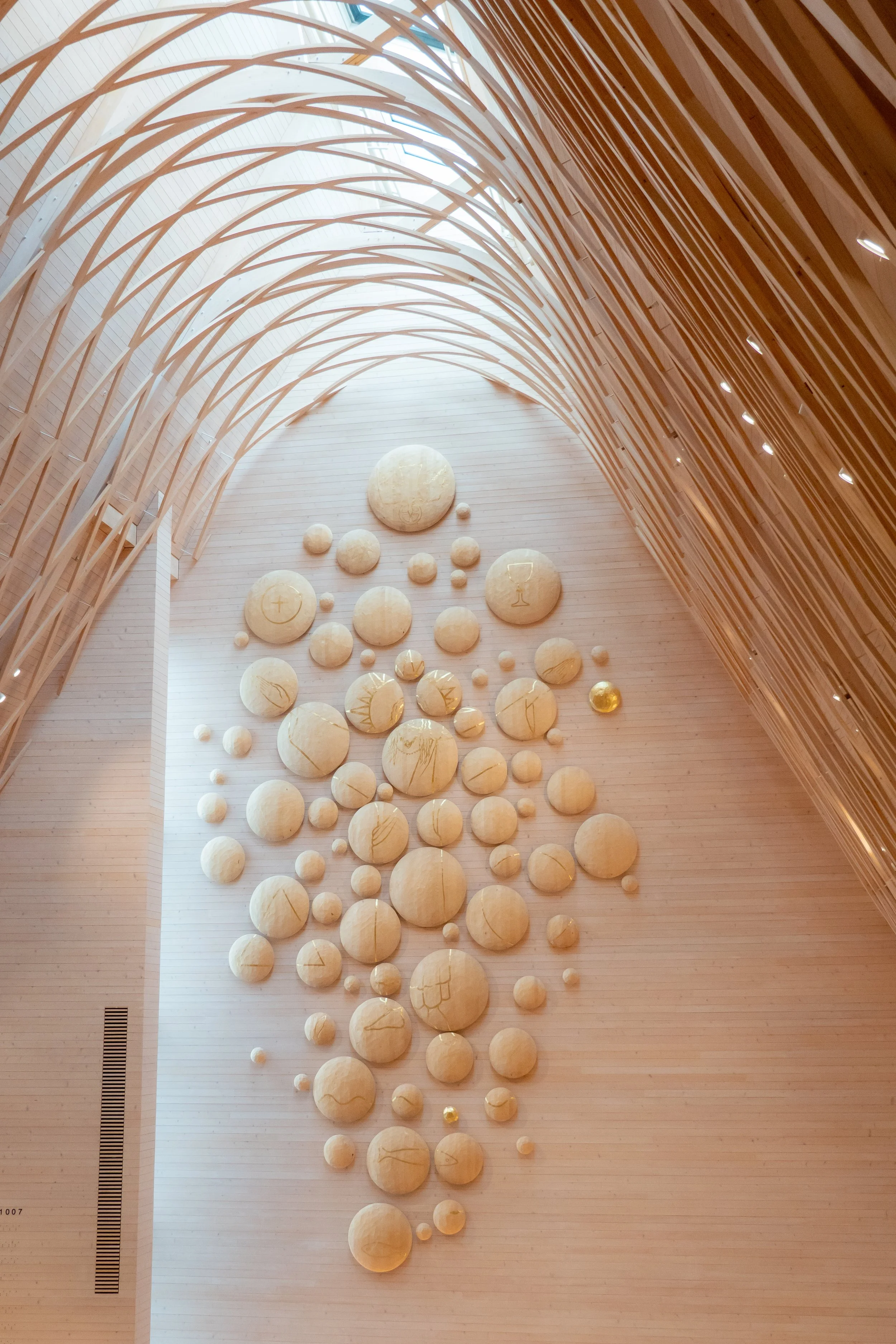 Interior view of a modern architectural space with a high ceiling and large wooden arches. Decorated with numerous large, beige, spherical sculptures on a wooden wall, with some gold accents and line art symbols. Natural light filters in from above.