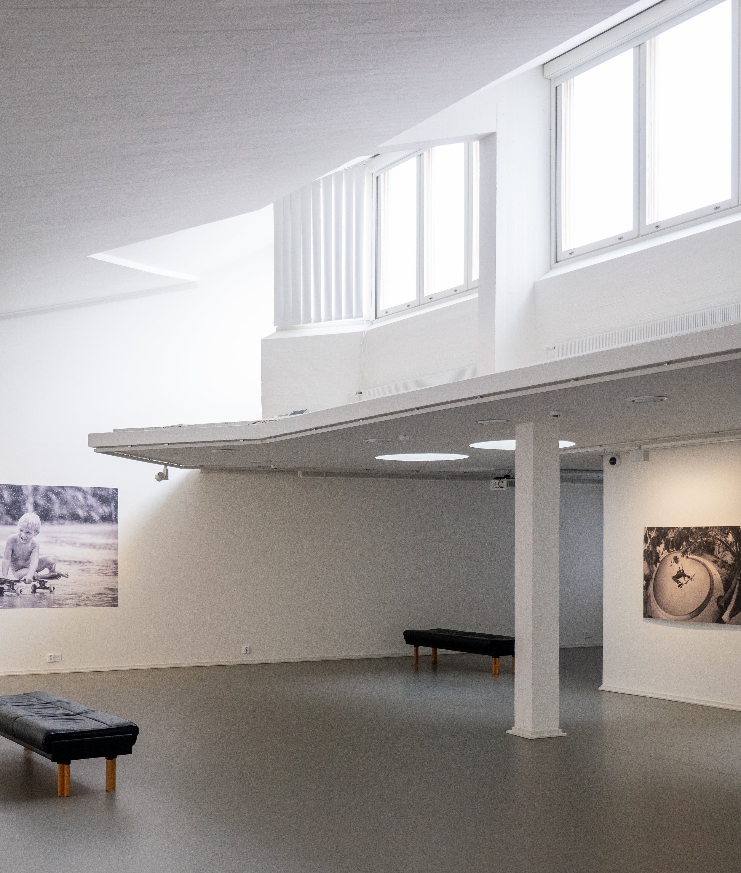 Interior of an art gallery with black and white photographs on white walls, benches, and large windows letting in natural light.