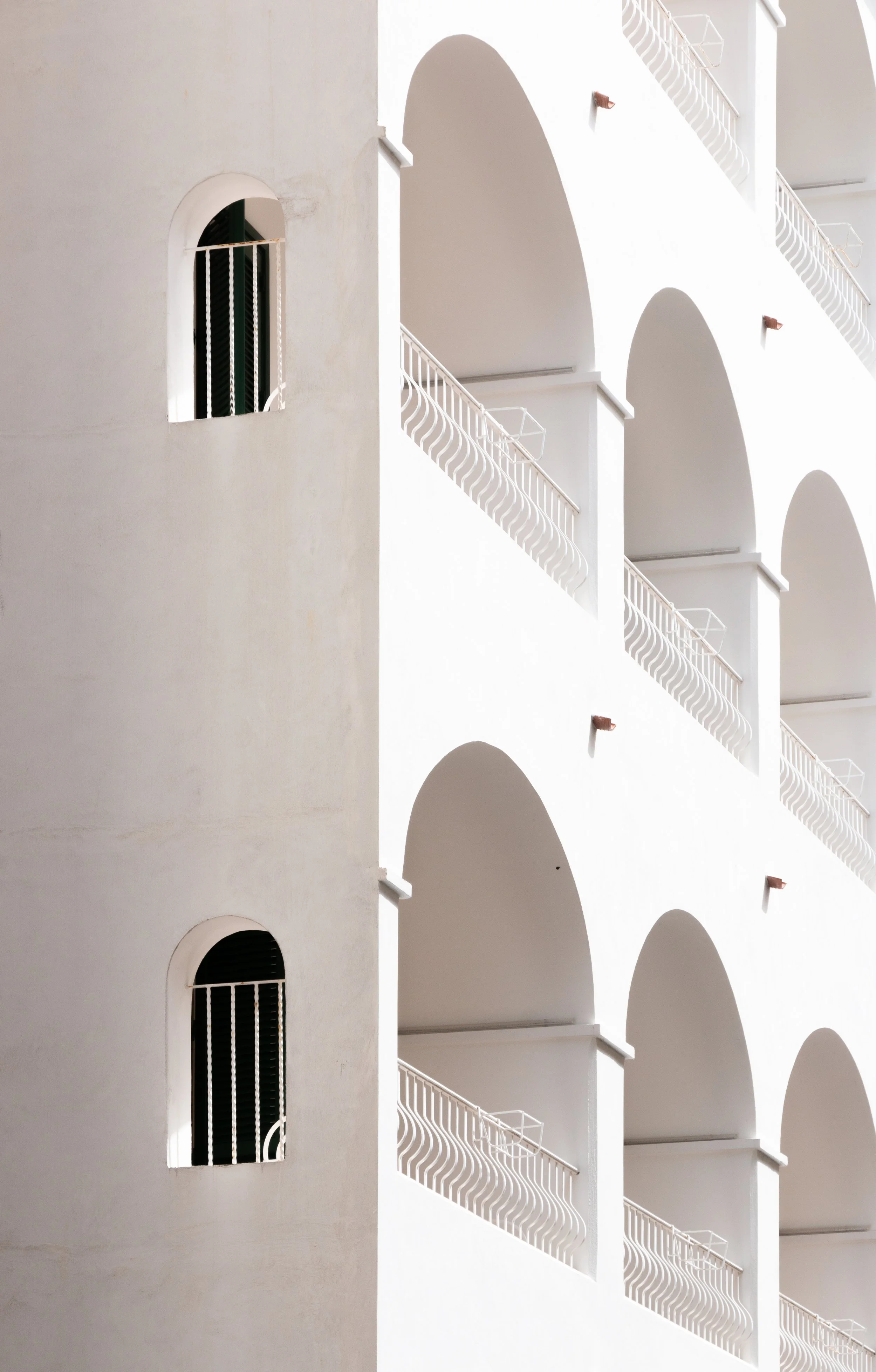 A building with white walls, featuring arched balconies with white railings and small windows with black bars.