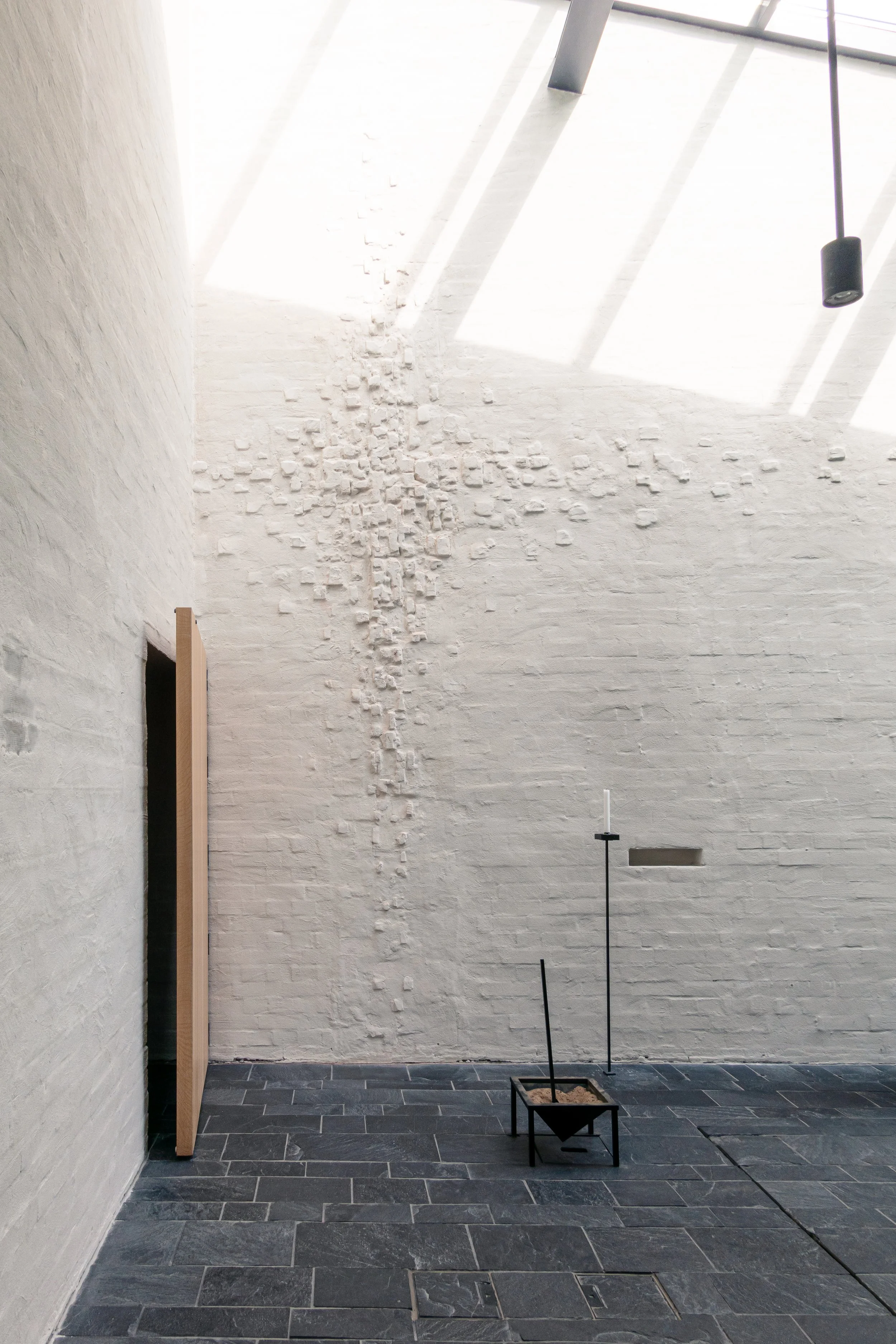 Minimalist interior with white brick wall, black tiled floor, and natural light from skylights. Art installation features a small black frame with a mound of sand, a black pole with a white candle, and a black horizontal shelf.