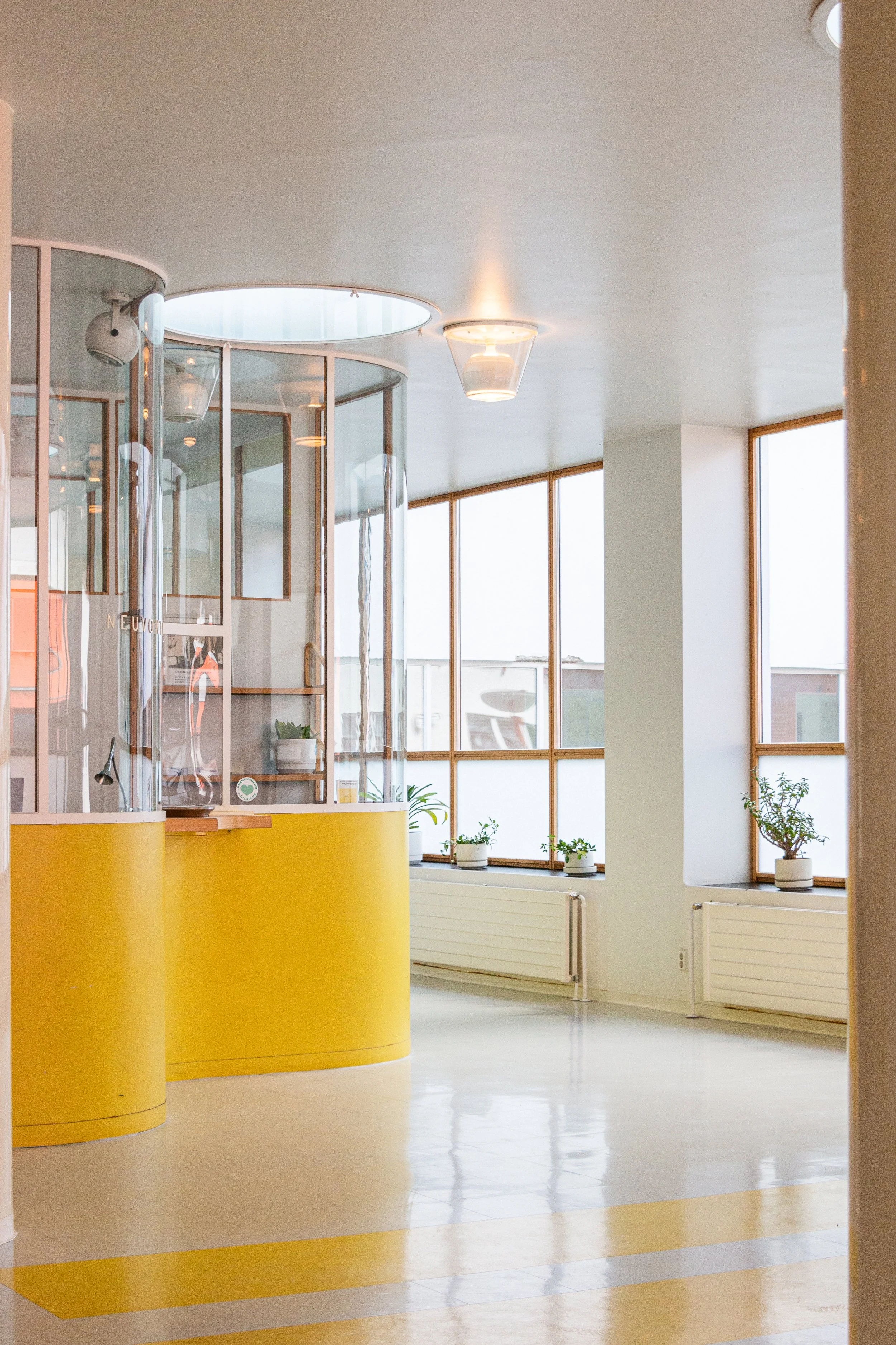 Interior of a modern, bright room with large windows and potted plants on the window sills. There is a yellow curved reception desk and a glass enclosure on the left side of the image.