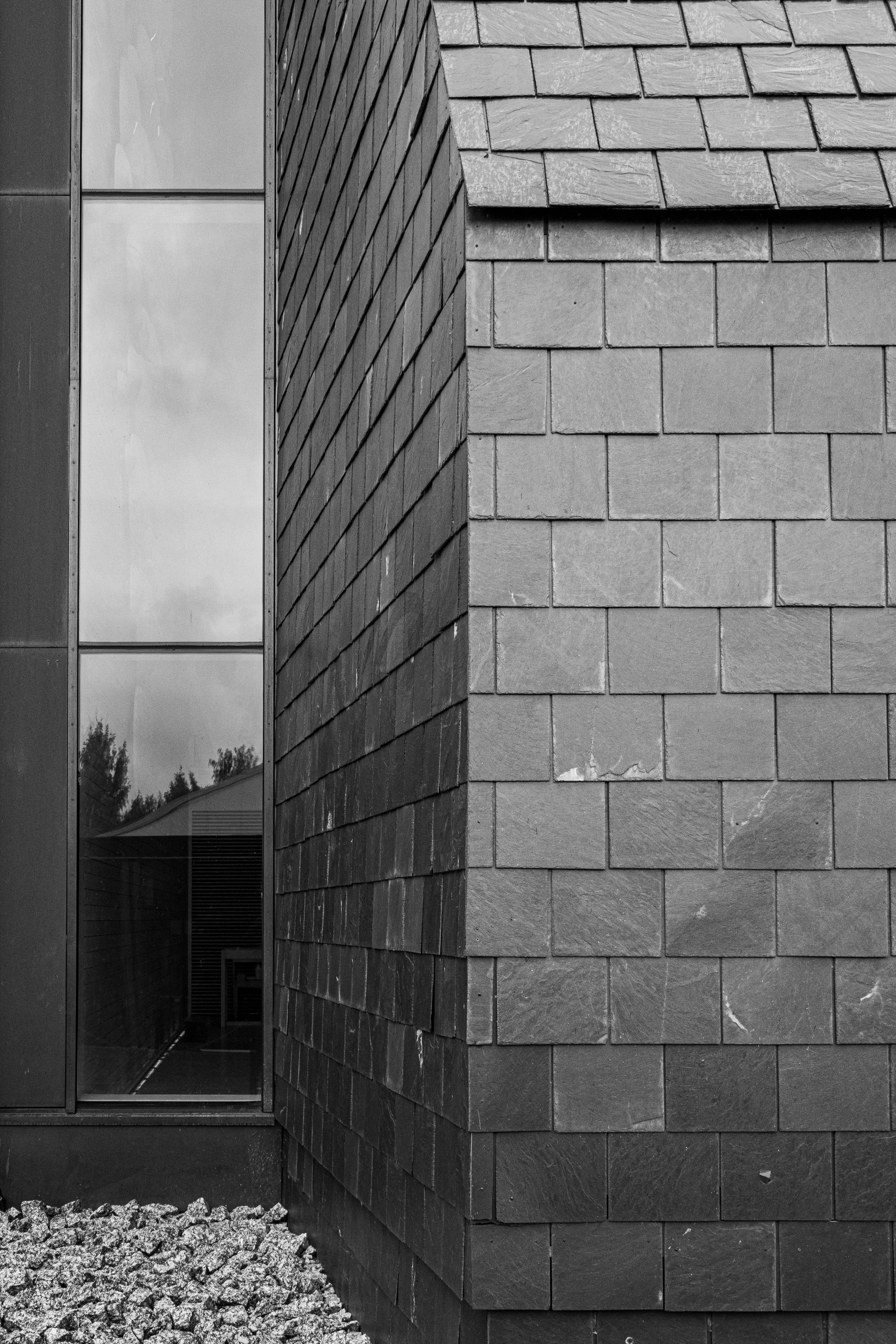 Close-up view of a building corner with brick and metal panels, and a window reflecting trees and a house roof.