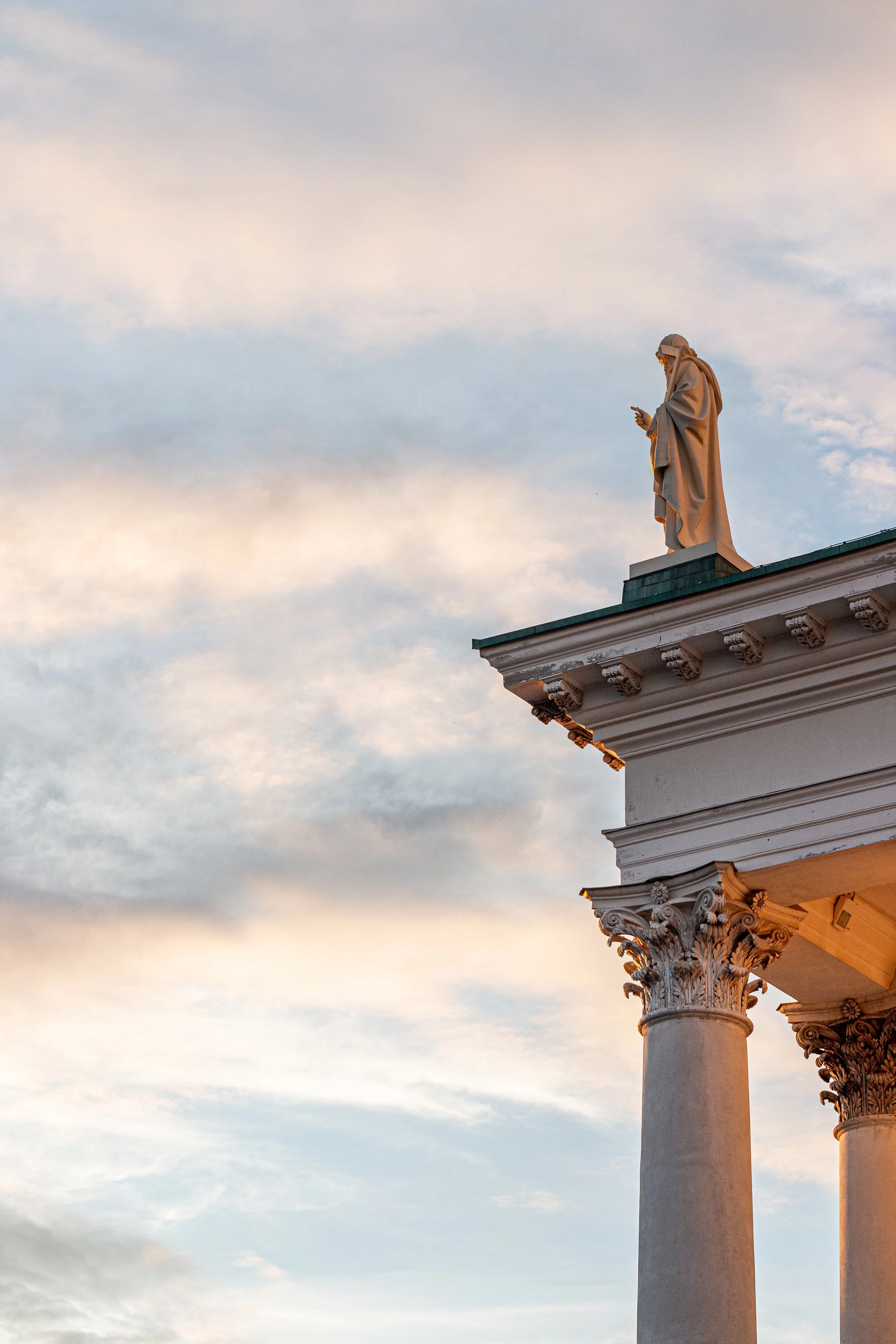 Statue of a man in a robe standing on a building ledge with ornate columns, looking at his phone, against a cloudy sky at sunset.