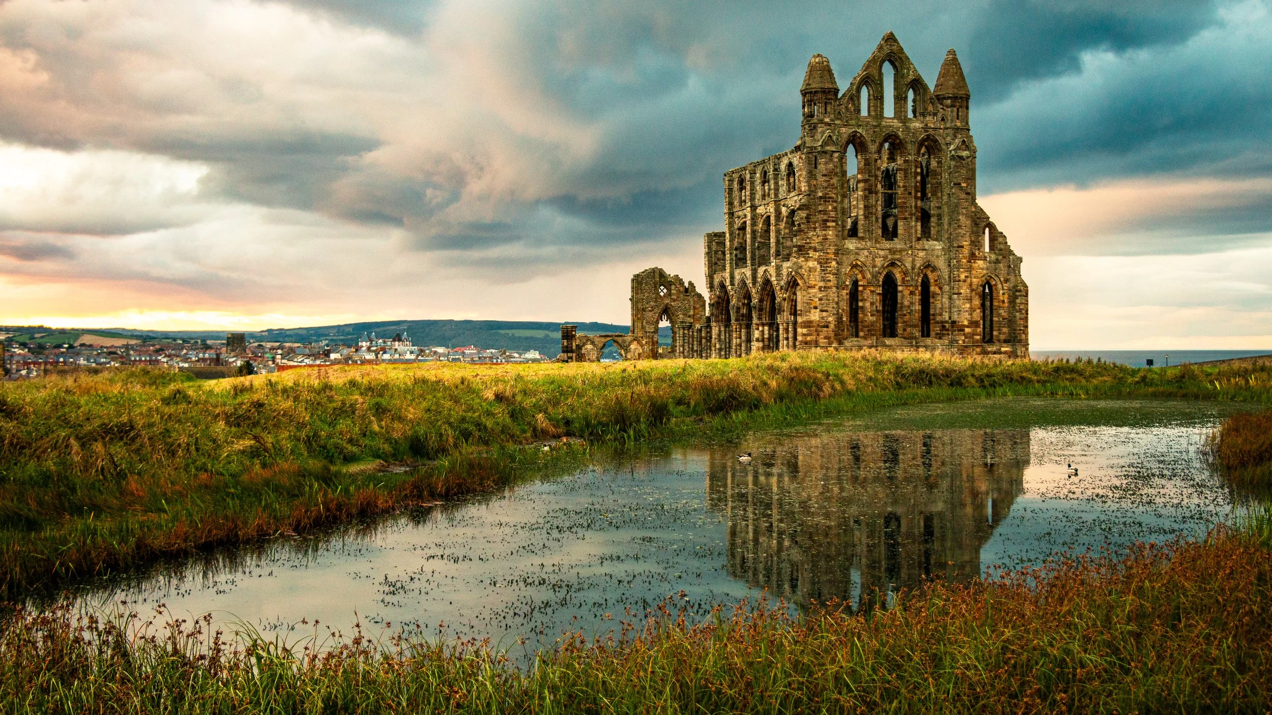 Ruins of an old stone church or castle with a pond reflecting the structure, grassy landscape, and a cloudy sky at sunset.