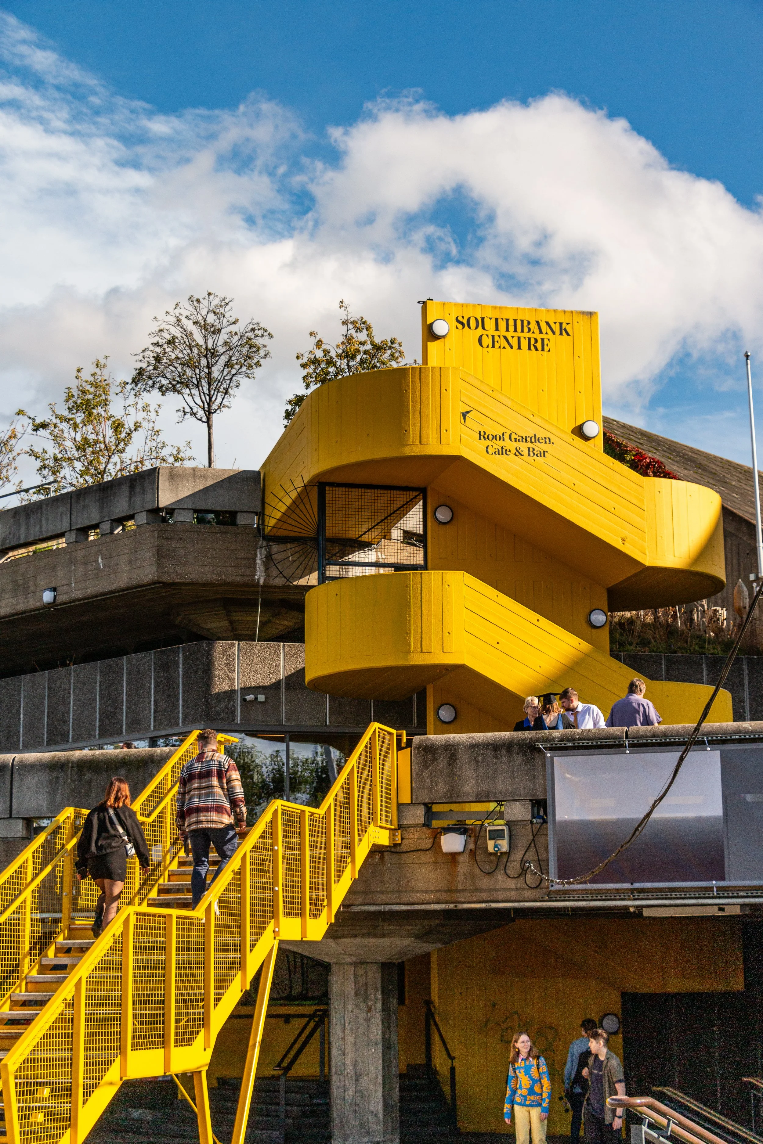 People walking up yellow stairs towards a bright yellow building labeled 'Southbank Centre' with additional signage for a roof garden, café, and bar. The building has a spiral yellow staircase outside, and the sky is partly cloudy.