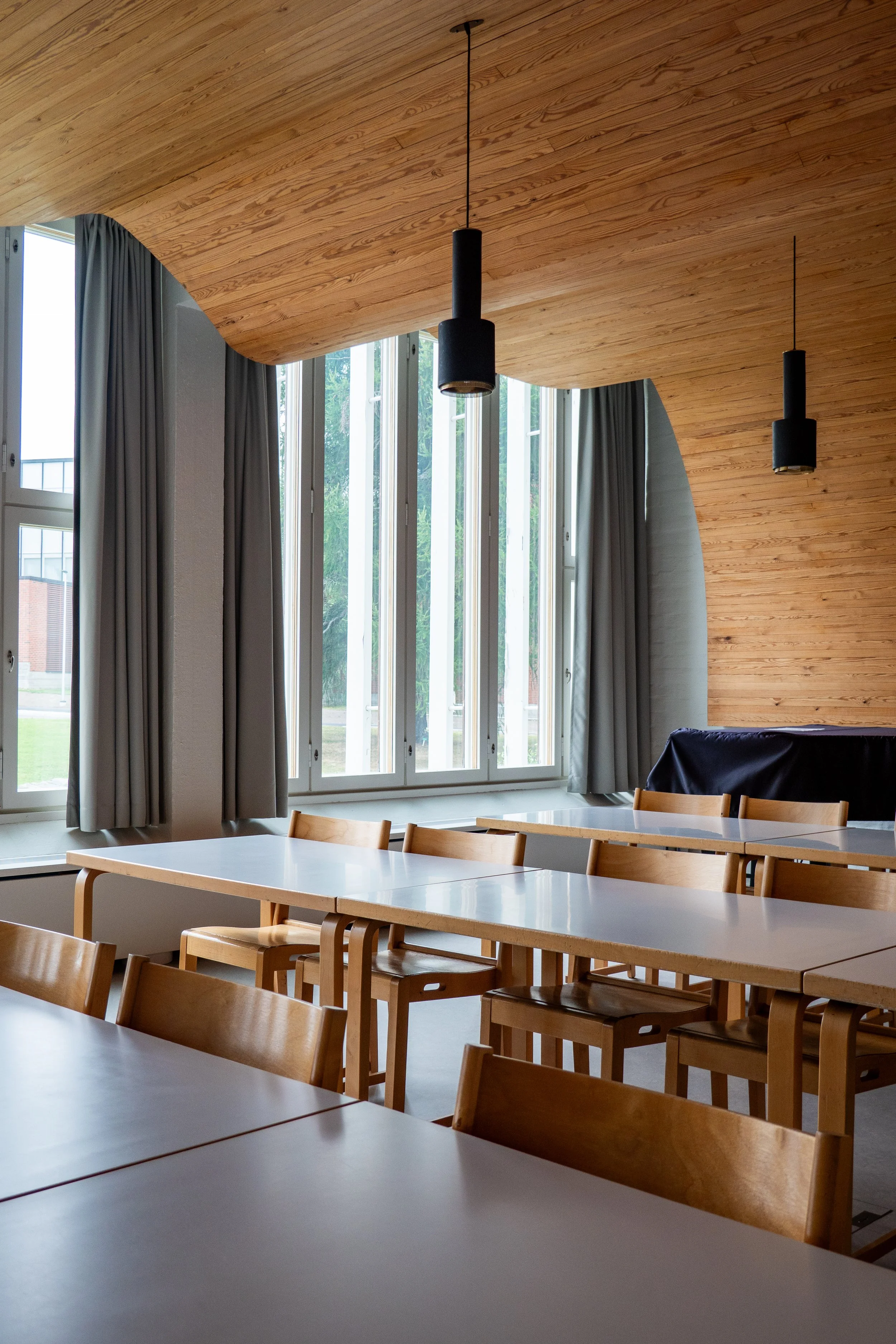 Empty classroom with wooden tables and chairs, large windows with gray curtains, and a curved wooden ceiling with two black pendant lights.