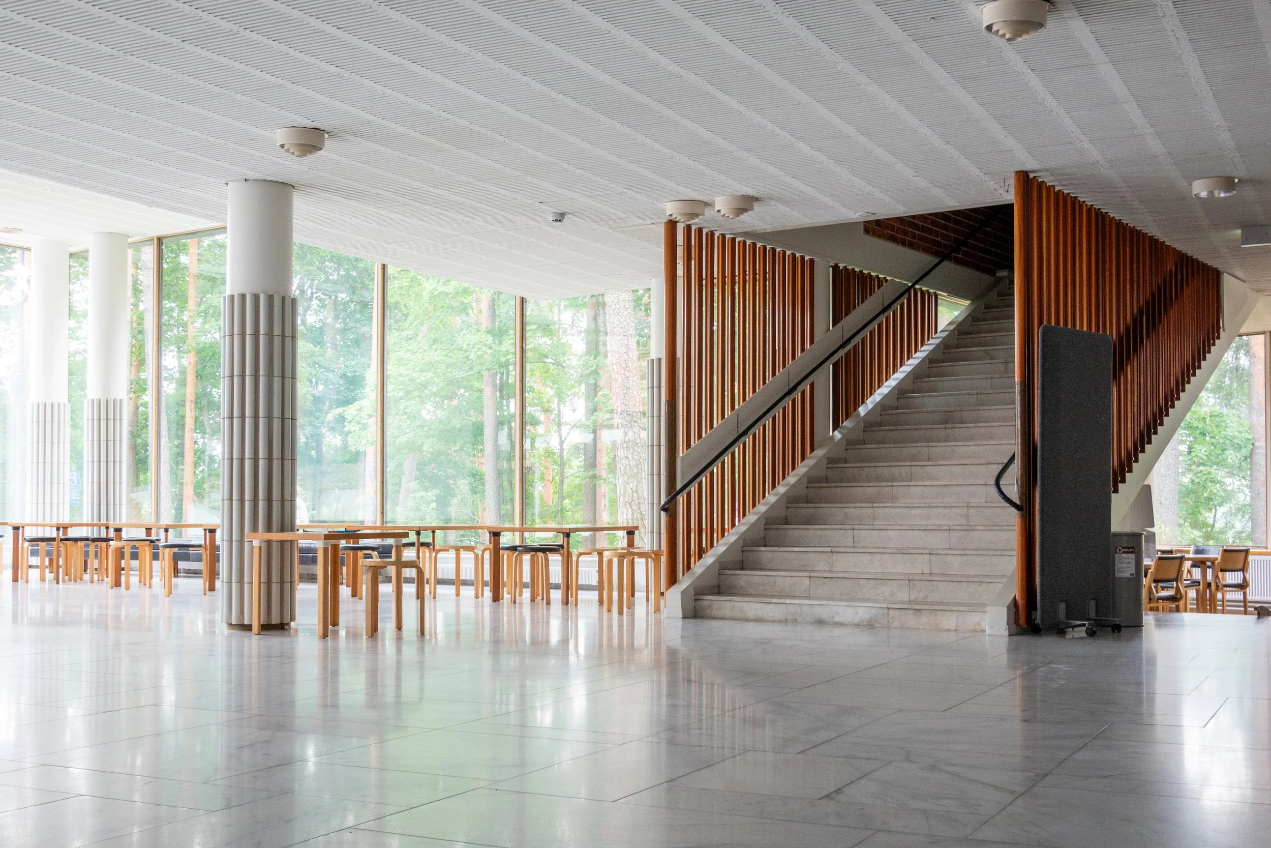 Interior of a modern building with large glass windows, wooden furniture, a staircase with wooden railing, and a view of green trees outside.