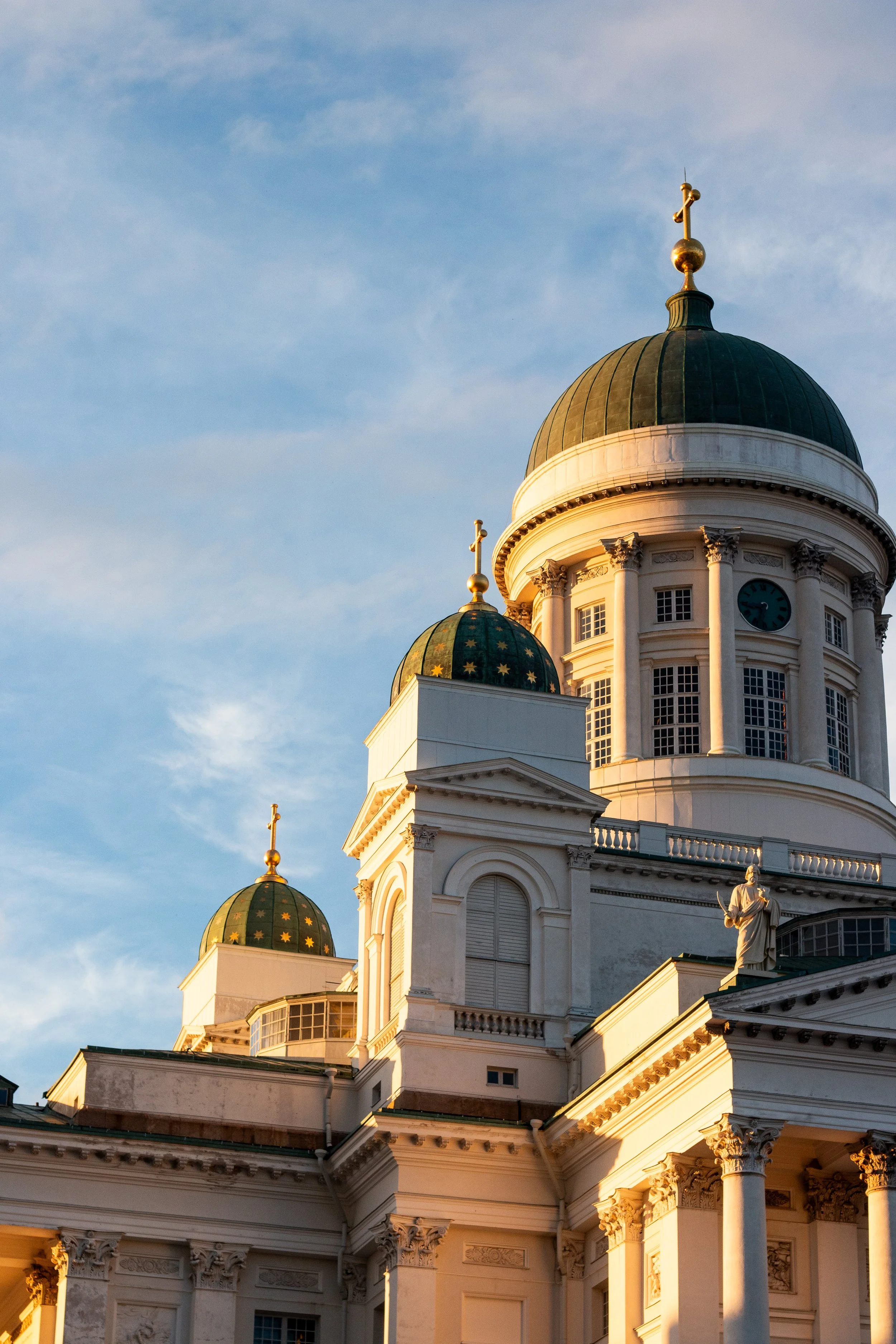 Photo of the large white and green-domed Helsinki cathedral with classical architectural features, statues, and a clock, captured during sunset or early evening.
