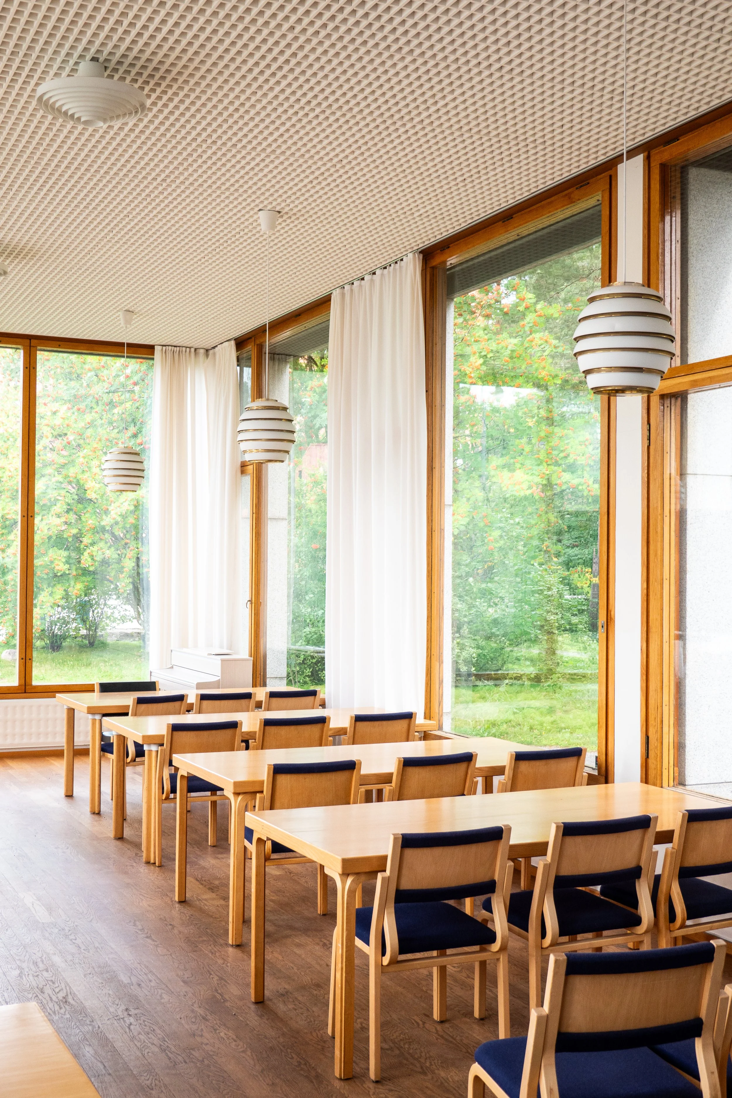 Interior of a bright, modern dining area with wooden tables and chairs with dark cushions, large windows with white curtains, and hanging pendant lights.