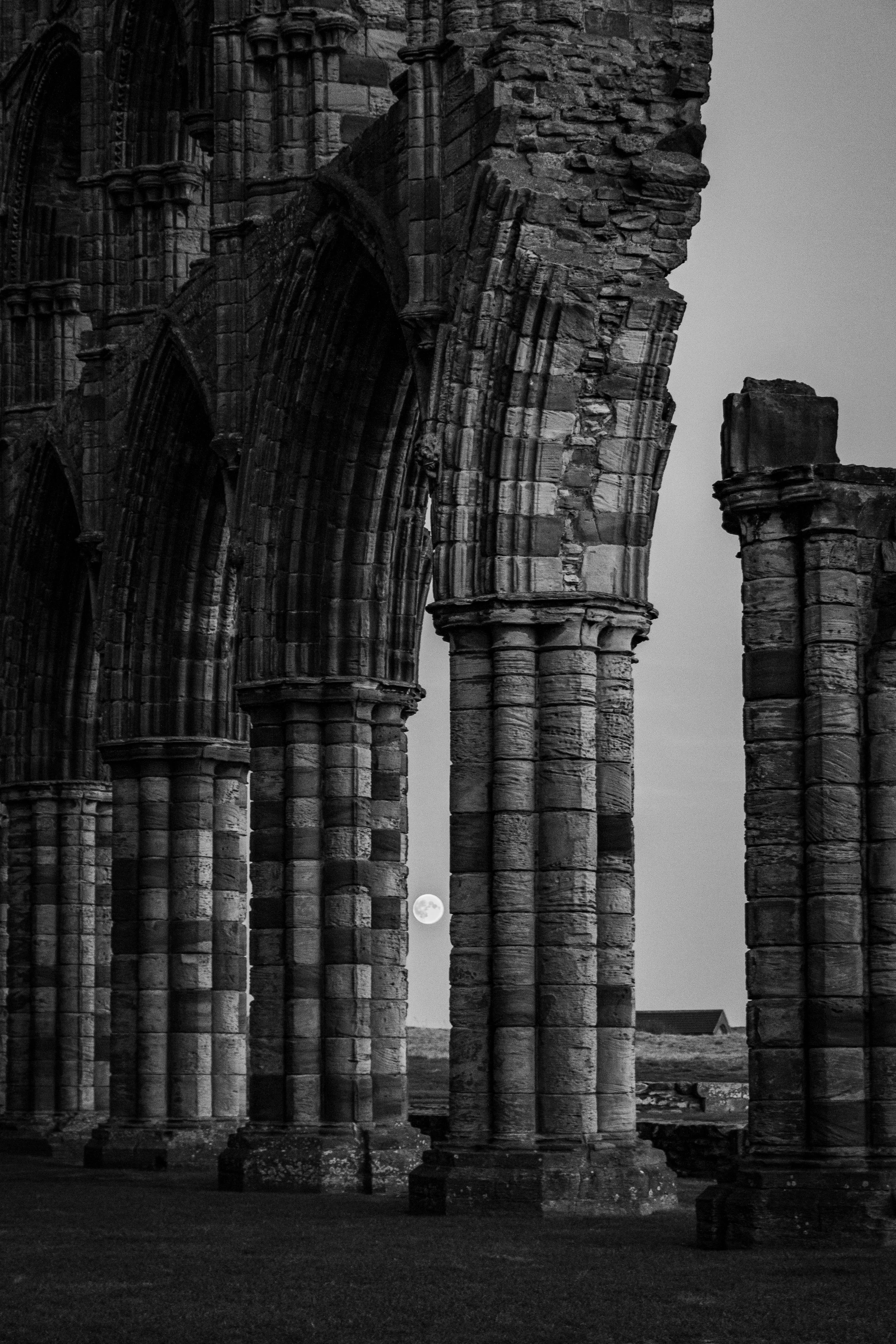 Black and white photo of ancient stone ruins of a Gothic cathedral with the moon visible between the columns.