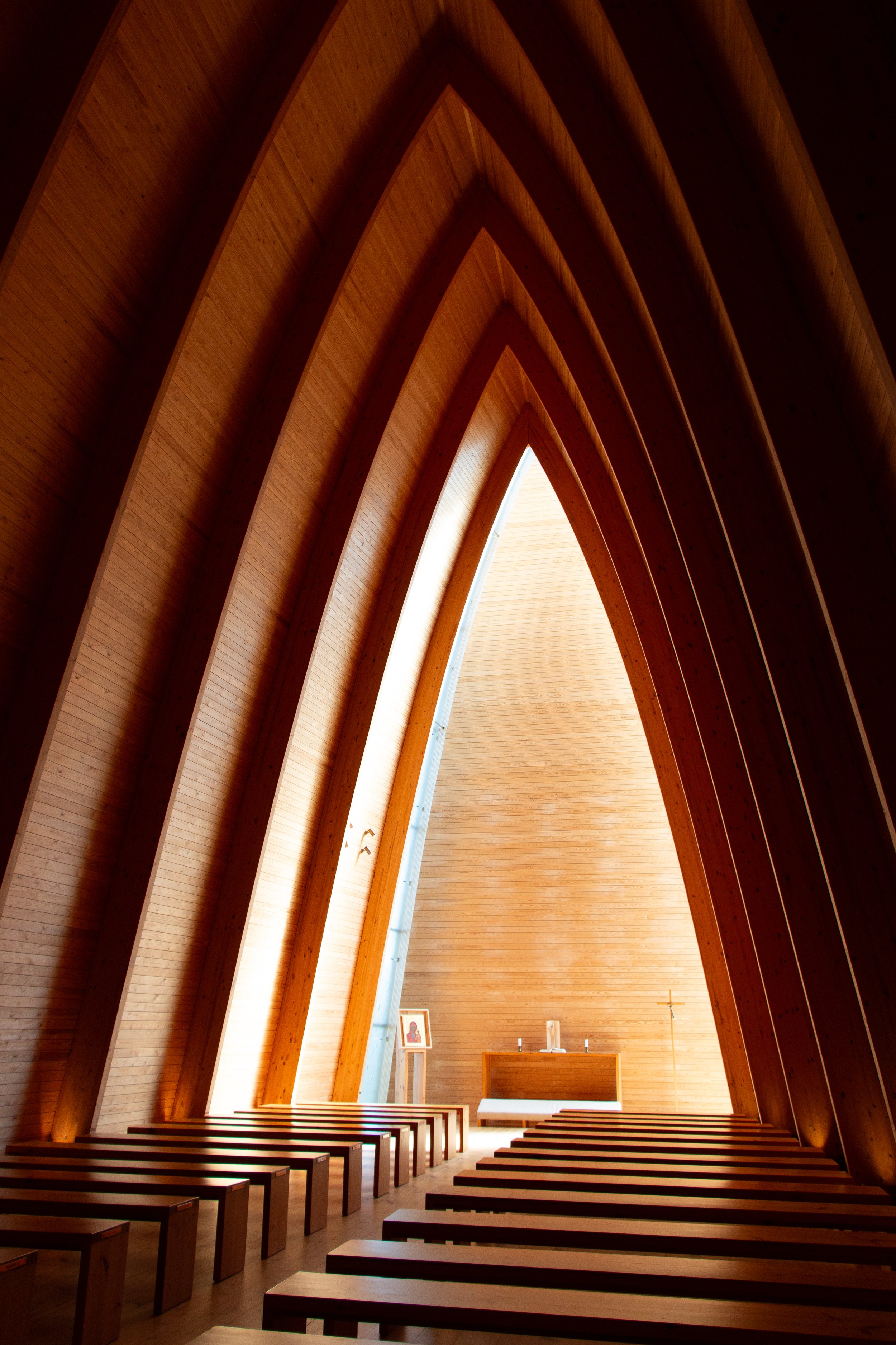 Interior of a modern wooden church with high, pointed arched ceilings, rows of benches, an altar at the front, and religious symbols including a cross and a framed picture.