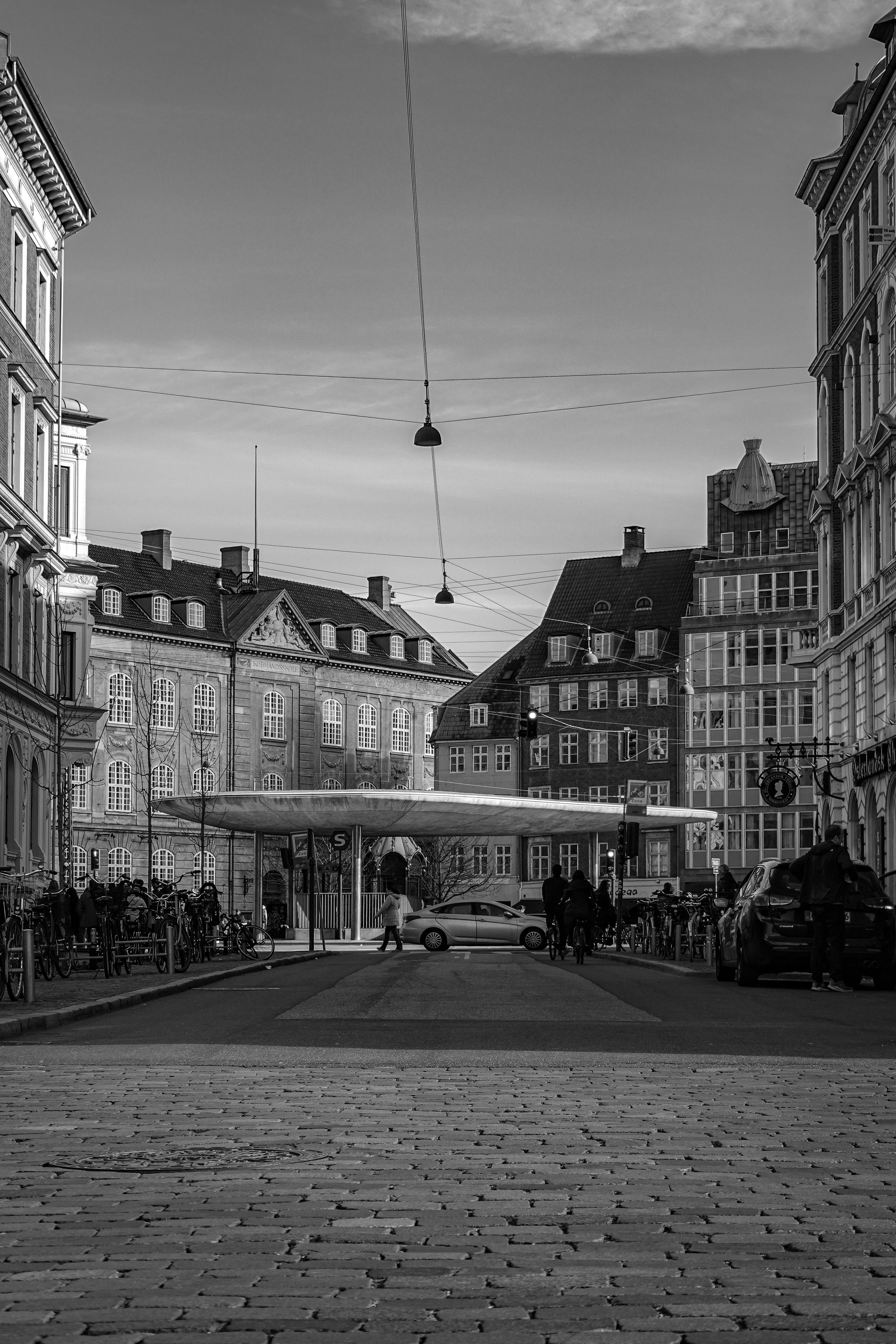 Black and white photo of a city street with old buildings, a modern bus stop canopy, parked bicycles, cars, and pedestrians, with tram wires overhead.