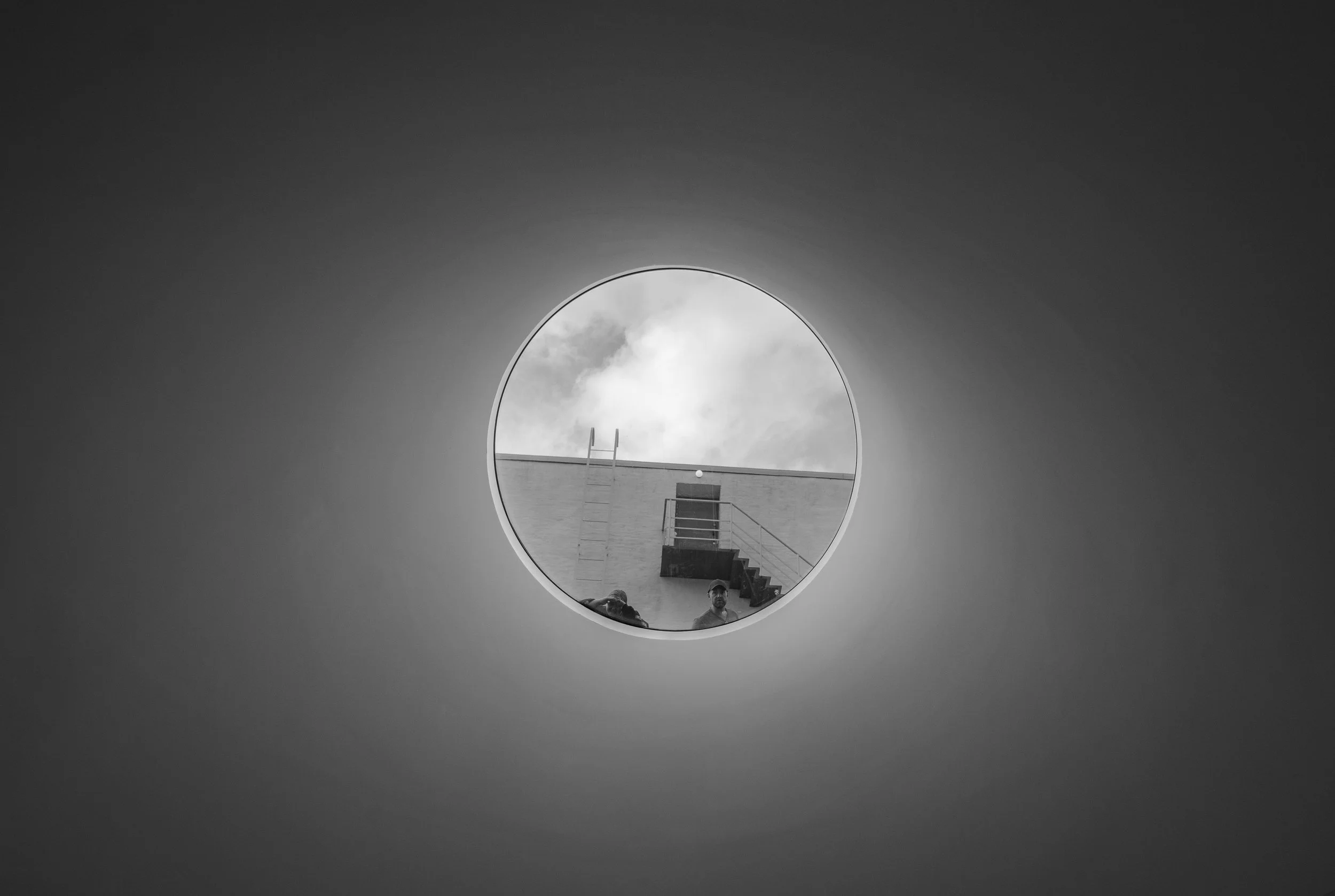 View through a round window showing part of a rooftop with stairs, a ladder, a railing, two people, and a cloudy sky in black and white.