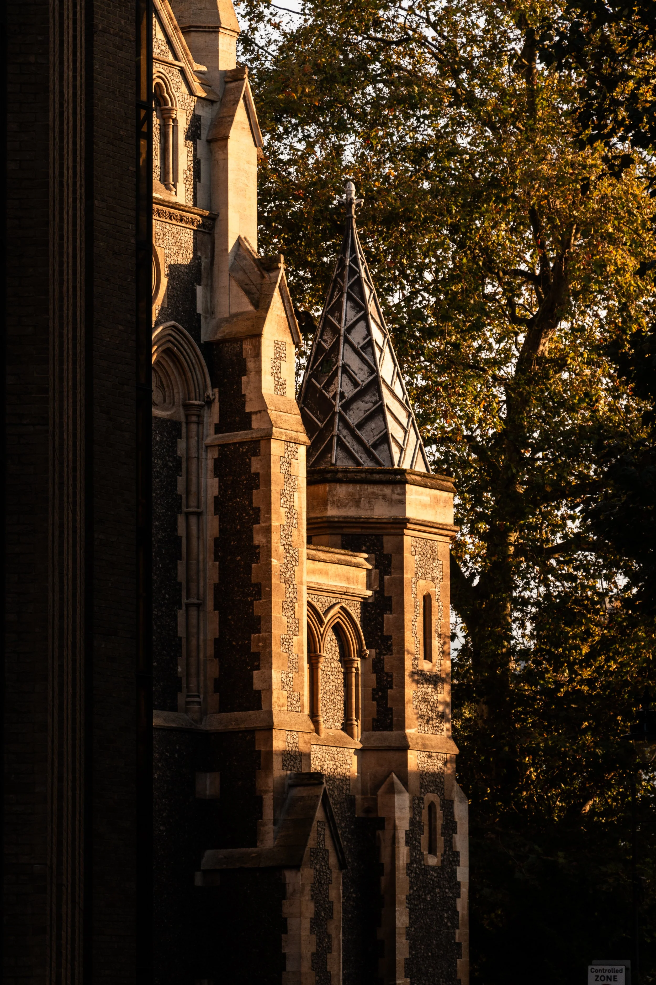 Close-up view of a historic church or castle with Gothic architectural features, including arched windows, decorative brickwork, and a pointed tower, illuminated by warm sunlight with trees in the background.