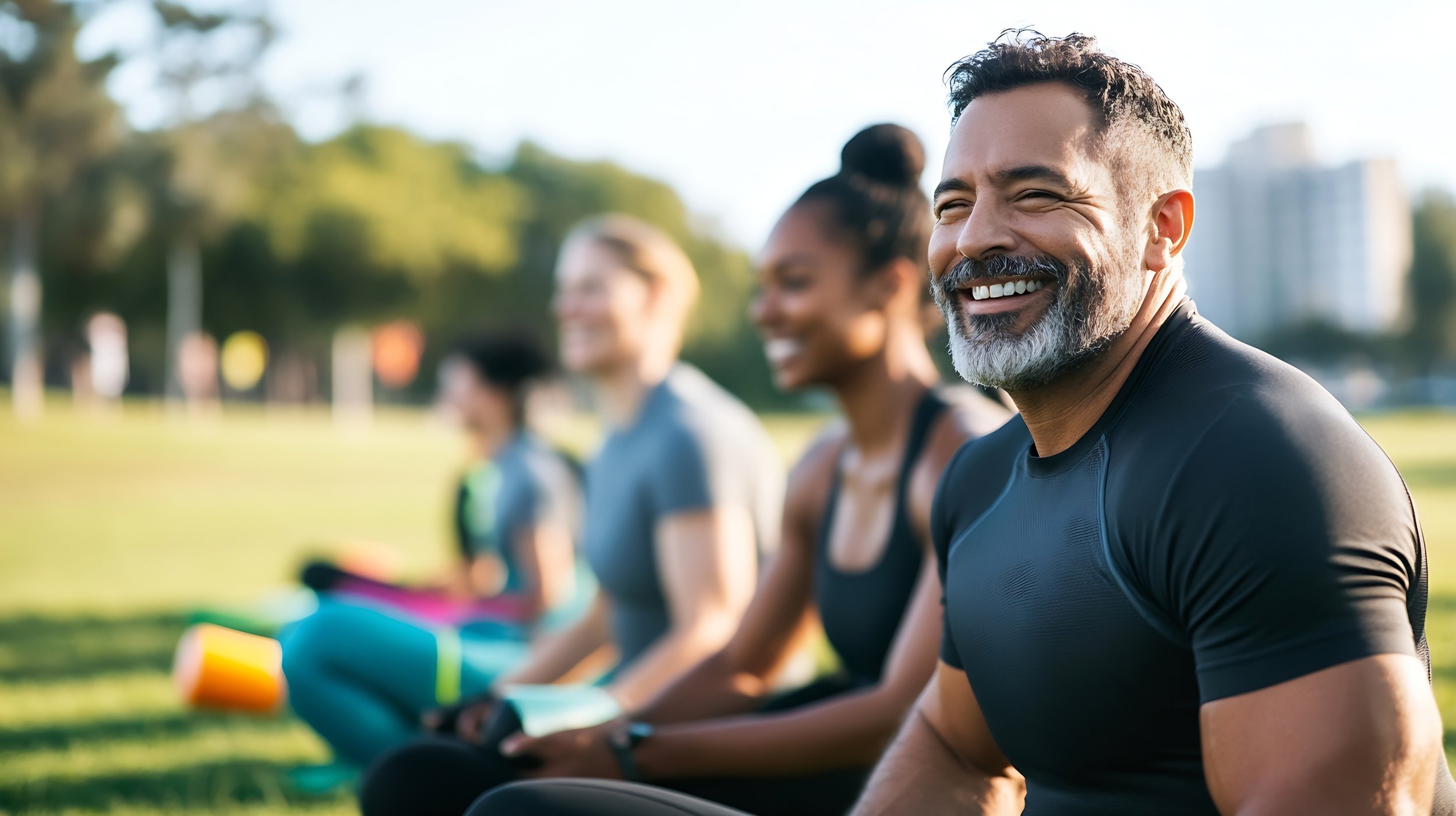 Group of diverse people sitting in a park, smiling, after exercise or yoga session, with the focus on a smiling man with a beard in the foreground.