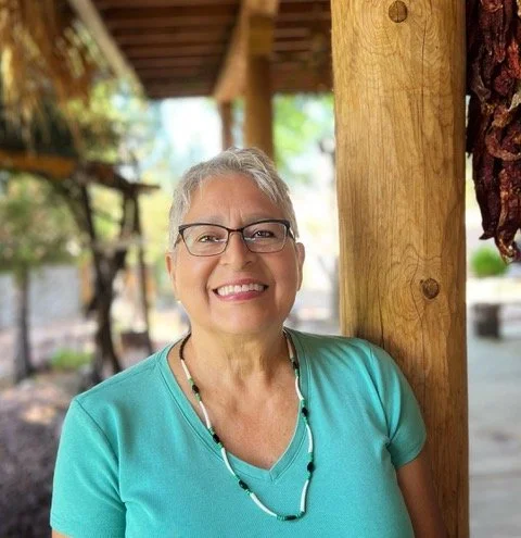 An elderly woman with short gray hair and glasses, smiling while standing outside near a wooden post in a bright, outdoor setting.