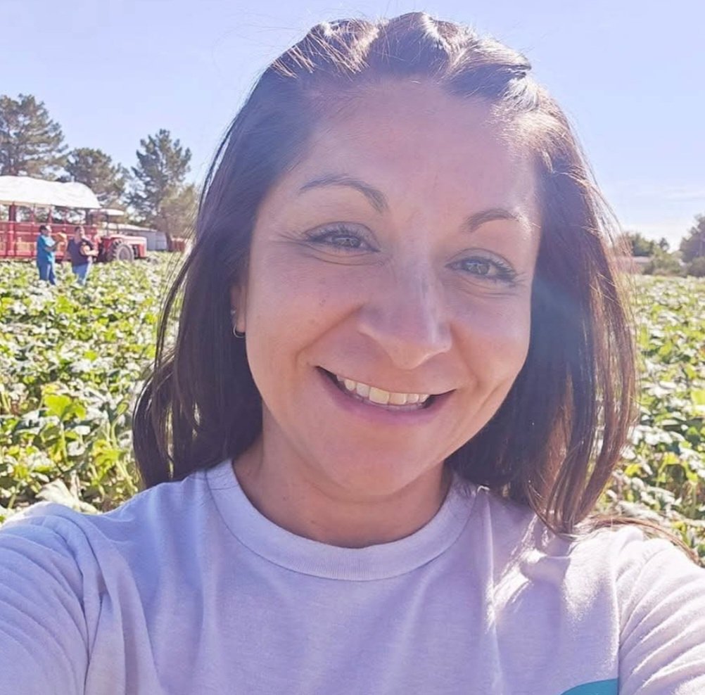 A woman with brown hair and blue eyes smiling outdoors in a sunny farm field with green crops. There are people and a red tractor in the background.