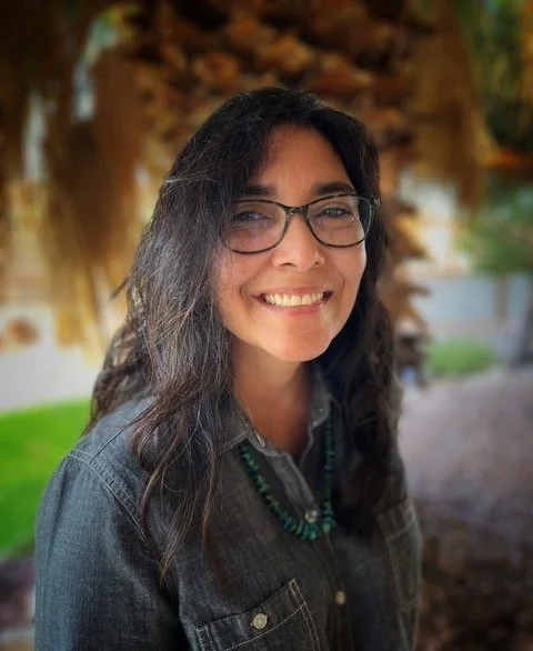 A woman with long dark hair and glasses smiling outdoors, with a blurred background of greenery and a tree.