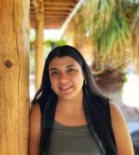 A young woman with long dark hair smiling outdoors, standing next to a wooden post with tropical plants in the background.