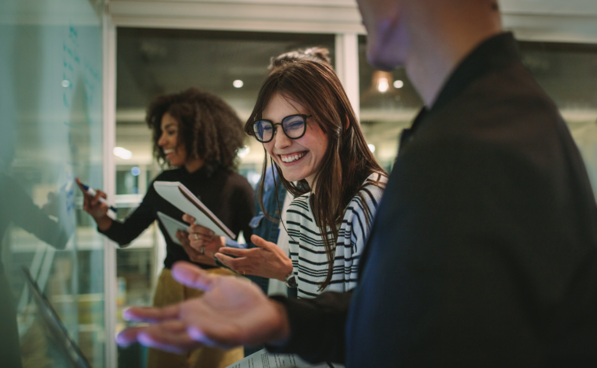 People collaborating and laughing in a professional meeting or workshop, standing around a whiteboard.