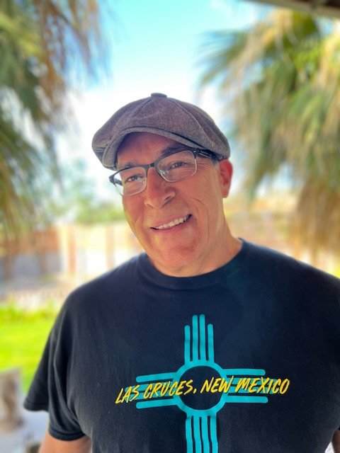 A smiling man wearing glasses, a gray cap, and a black T-shirt with a blue and yellow design and the text 'Las Cruces, New Mexico', outdoors with palm trees in the background.