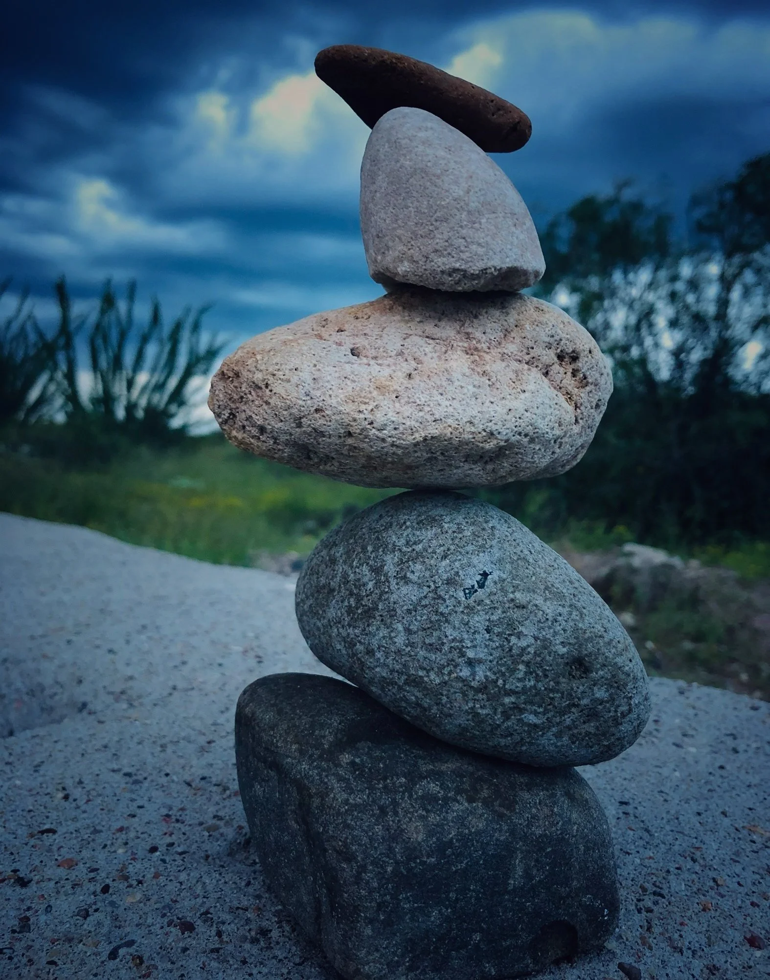 A stack of five balanced stones on sandy ground with a cloudy sky and some greenery in the background.
