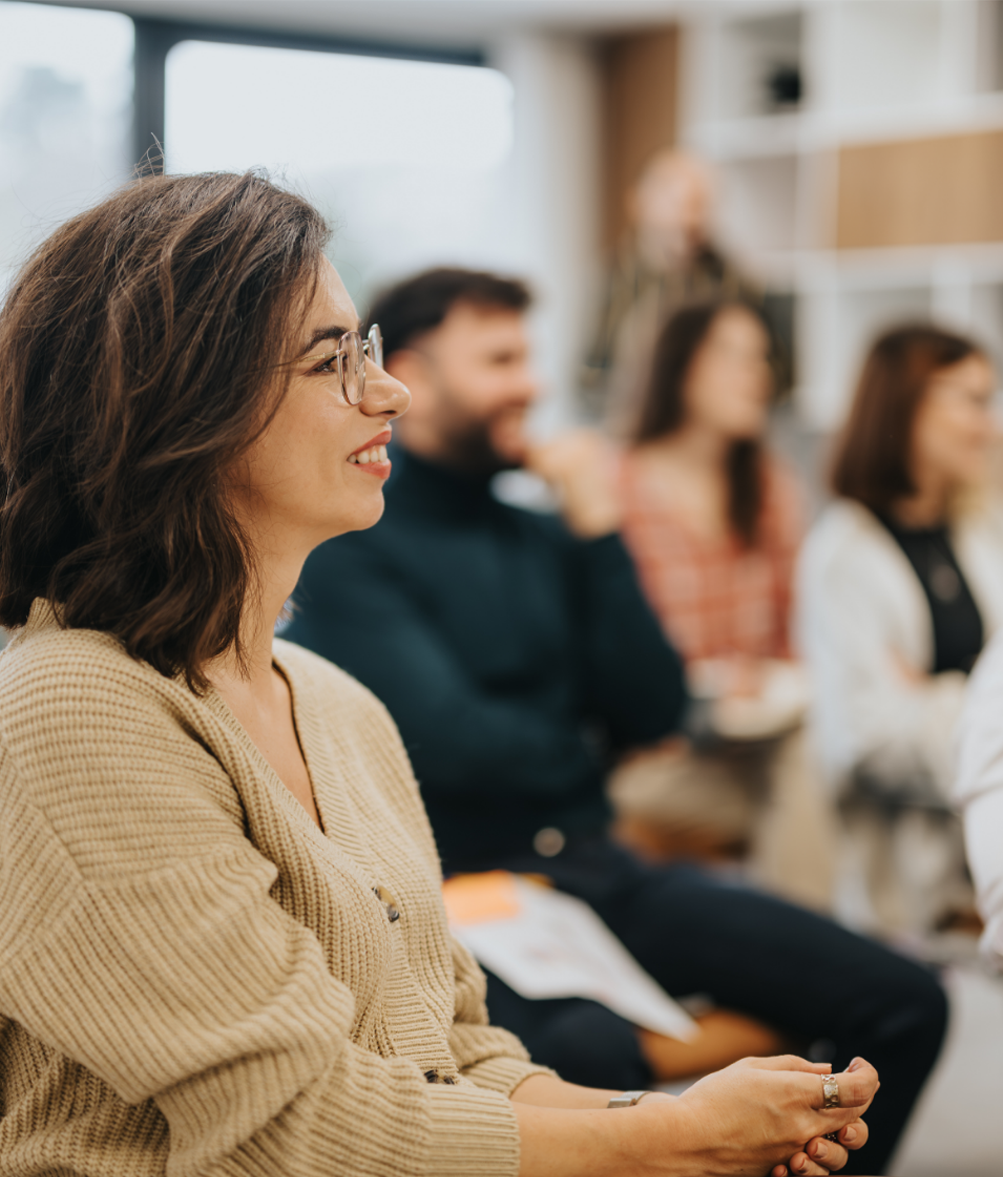 A woman with glasses and brown hair smiling, sitting at an event with other attendees in the background.