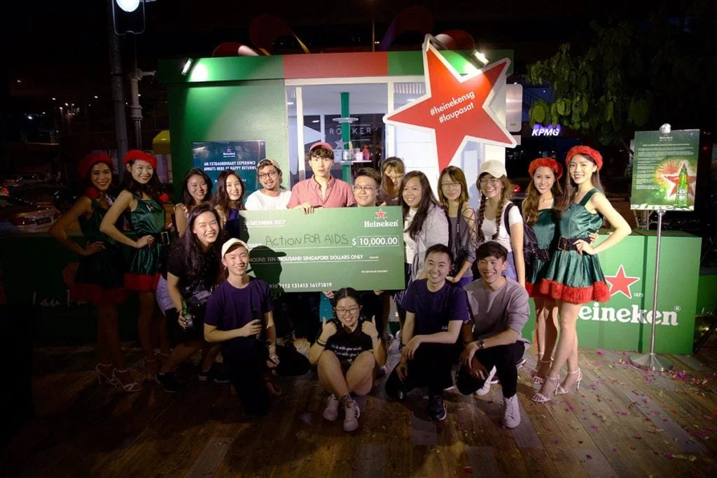 Group of people at a Heineken event holding a large mock check for $10,000 for AIDS charity, standing in front of a green and red Heineken-themed backdrop at night.