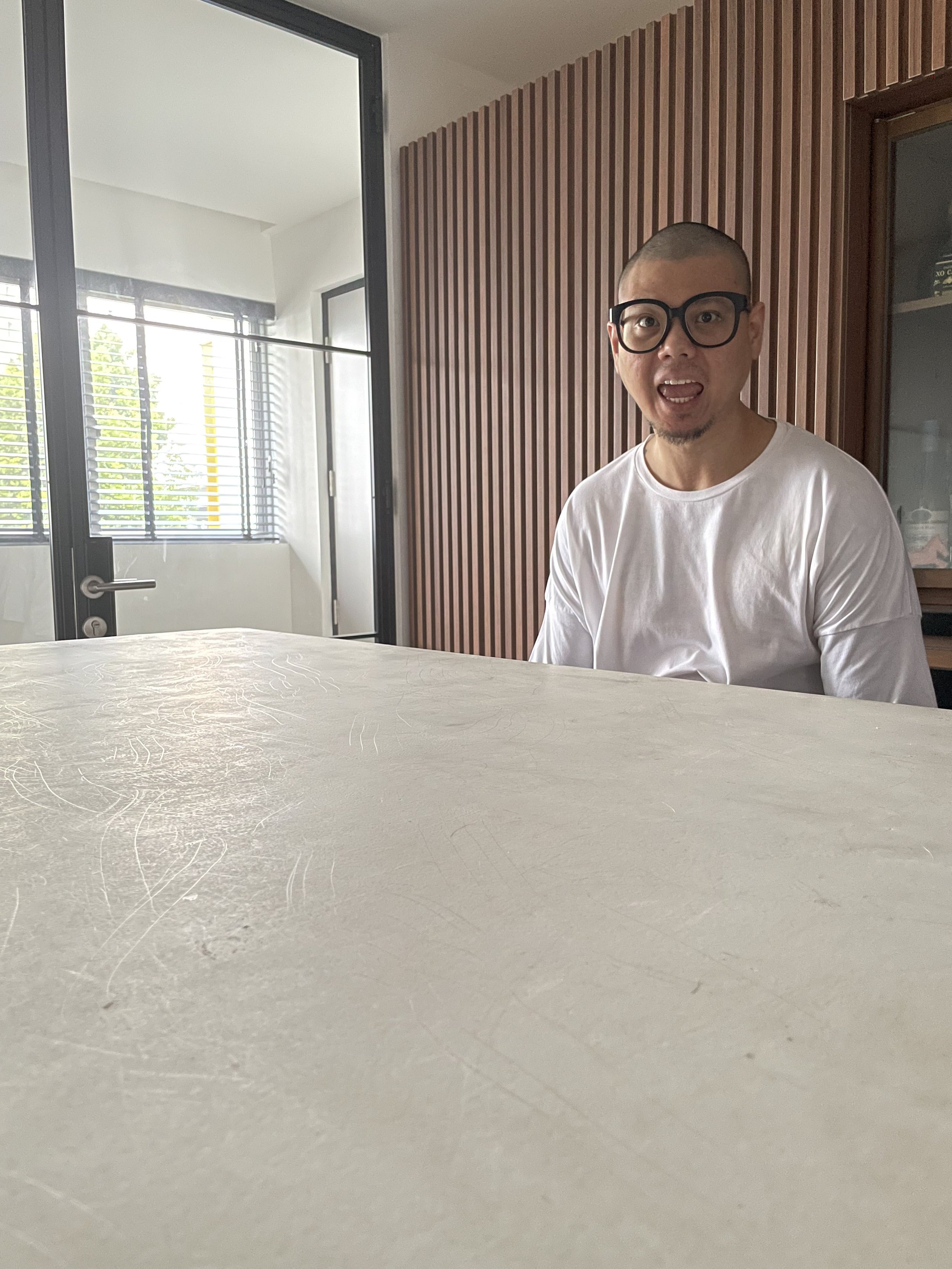 A man with glasses and a buzz cut, wearing a white shirt, sitting at a table inside a modern room with wooden paneling and a glass door leading outside.
