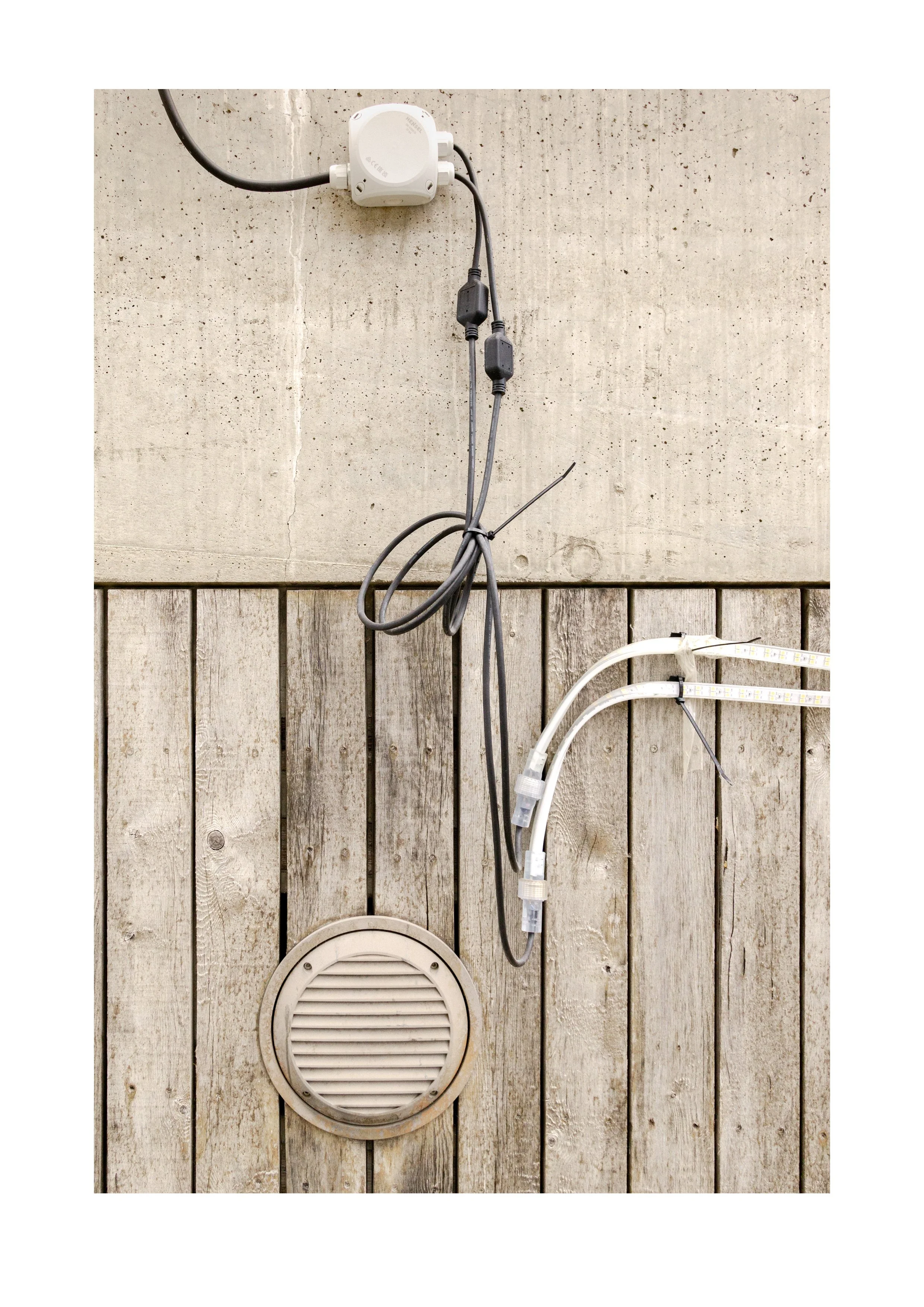 Photograph by Erik Nordby of electrical junction box with hanging cables and LED light strips above a circular wall vent on a wooden panel wall. Archive number 004812.