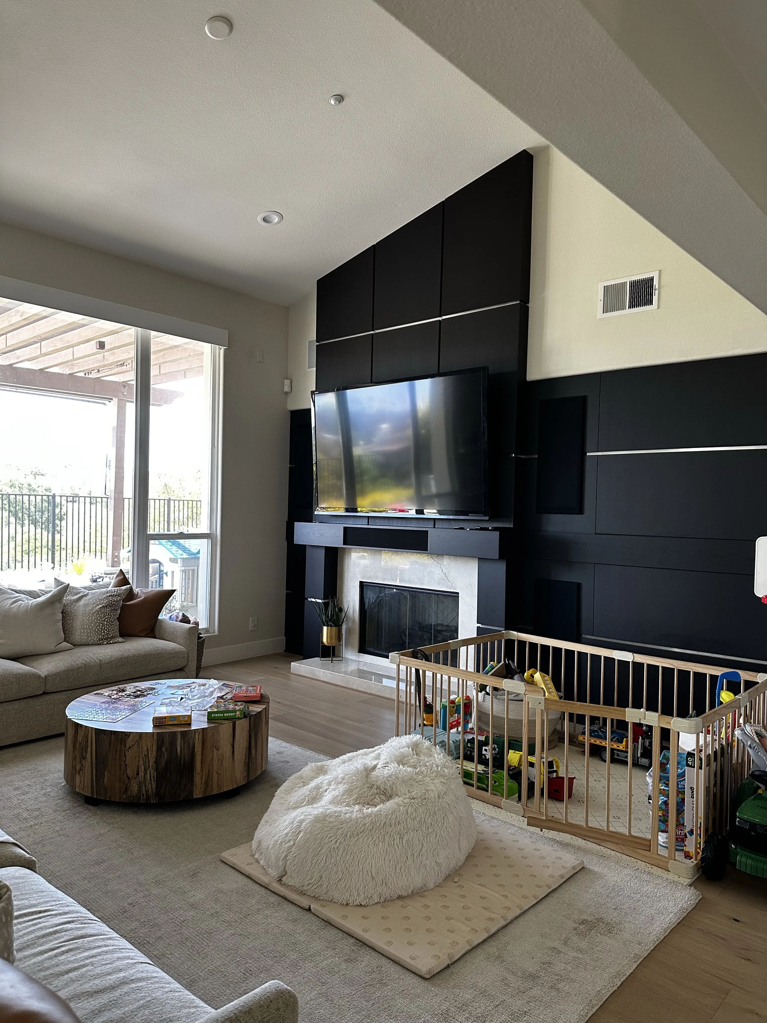 Living room with a beige couch, a round wooden coffee table, a fireplace, a flat-screen TV, a beige fluffy bean bag, and a playpen with children's toys near a large window and sliding glass door.