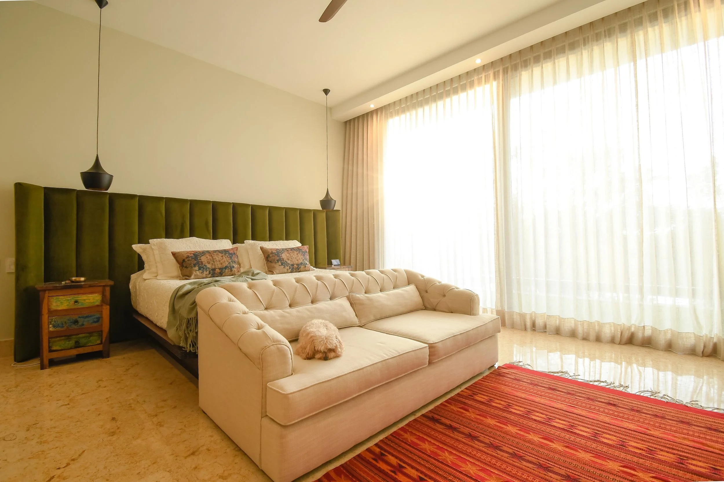 A bedroom with a large window covered by sheer curtains, a bed with a green velvet headboard, a beige tufted couch in front of the bed with a small dog lying on it, a red striped rug on the floor, and pendant lights hanging from the ceiling.