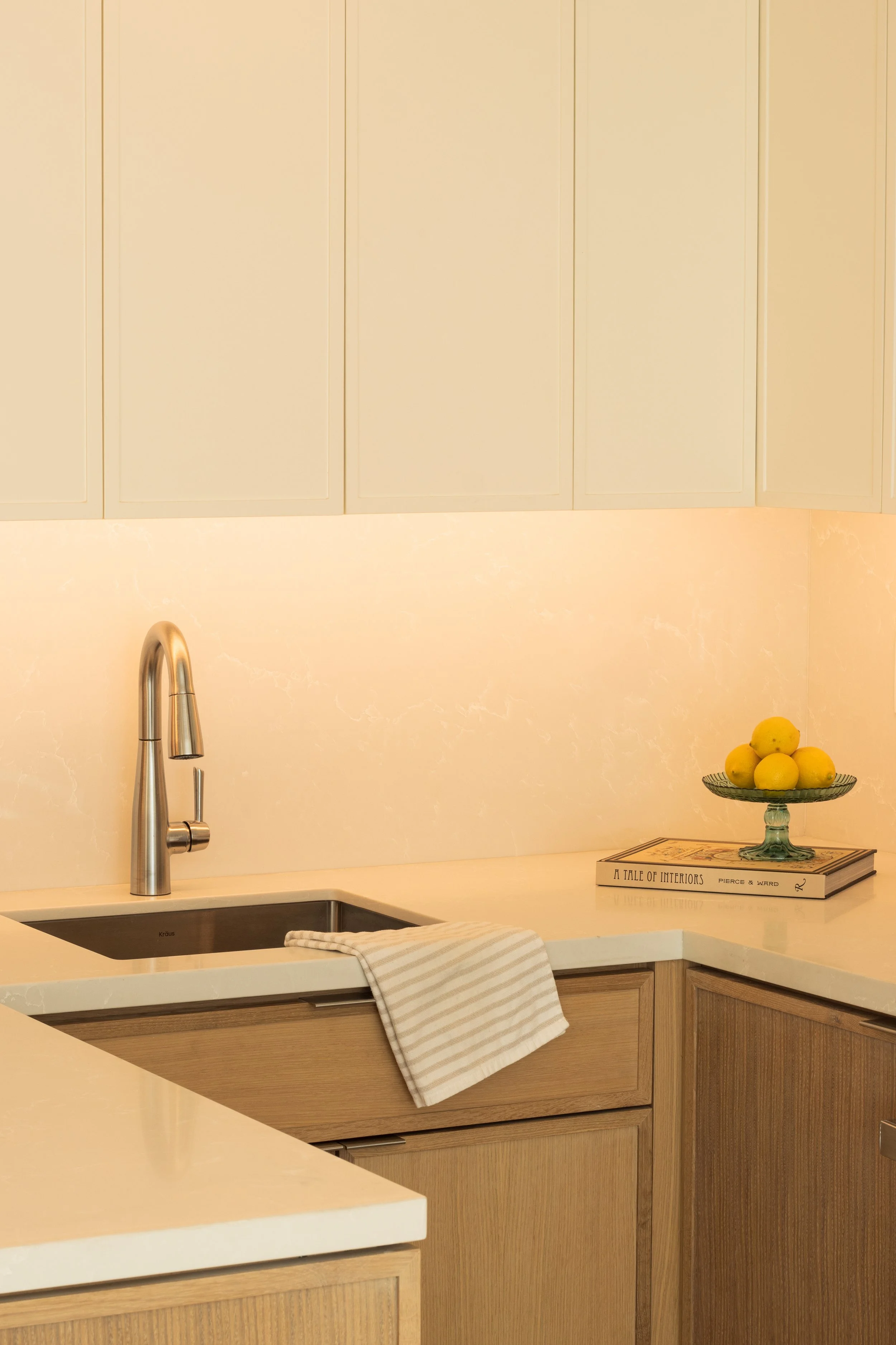 A modern kitchen with a beige backsplash, wooden cabinets, a stainless steel faucet, a white countertop with a dish towel, and a glass plate of lemons on top of a couple of books.