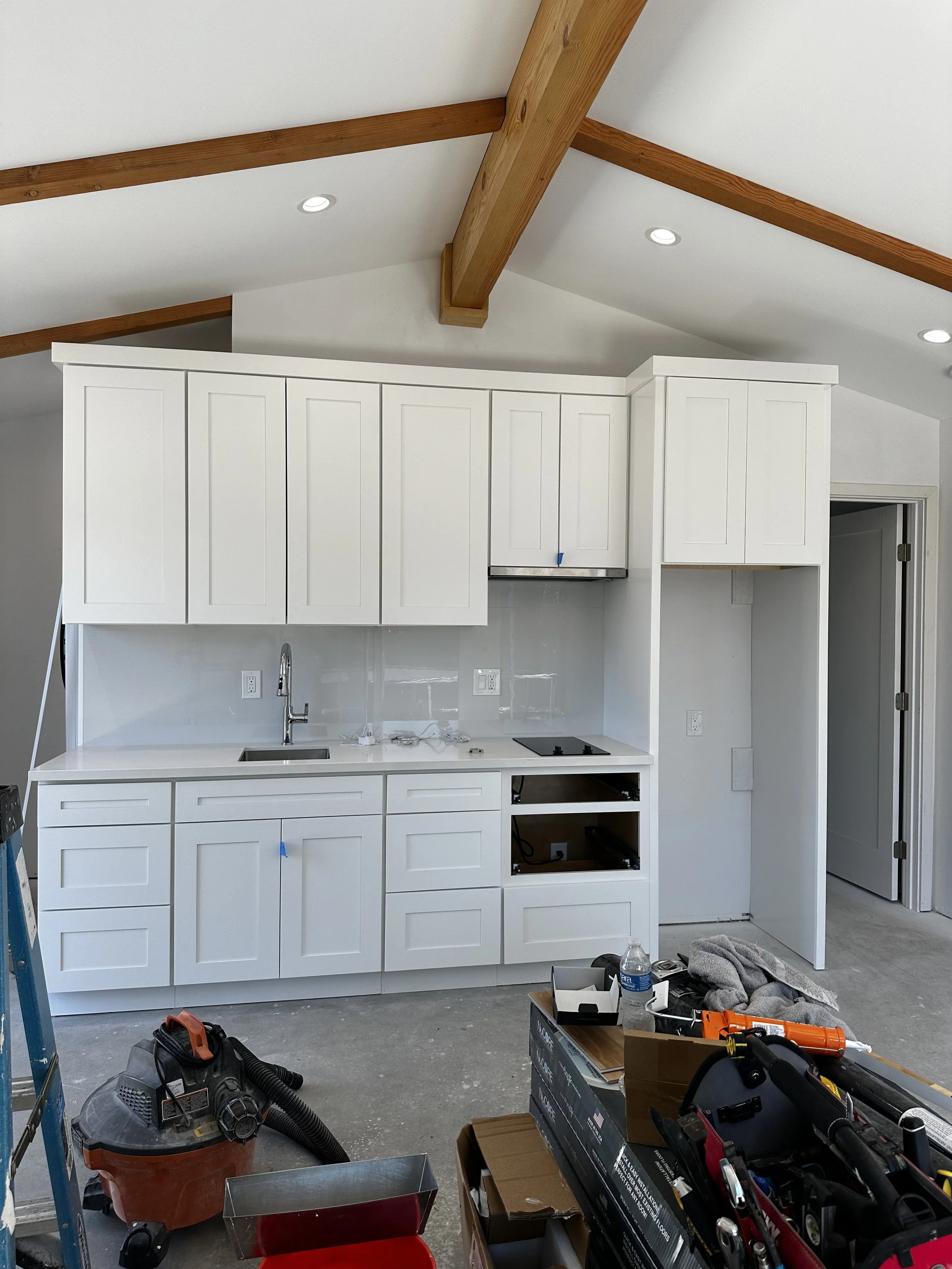 Currently under construction, a small kitchen with white cabinets, a sink, and a stove, with tools and materials on the floor.