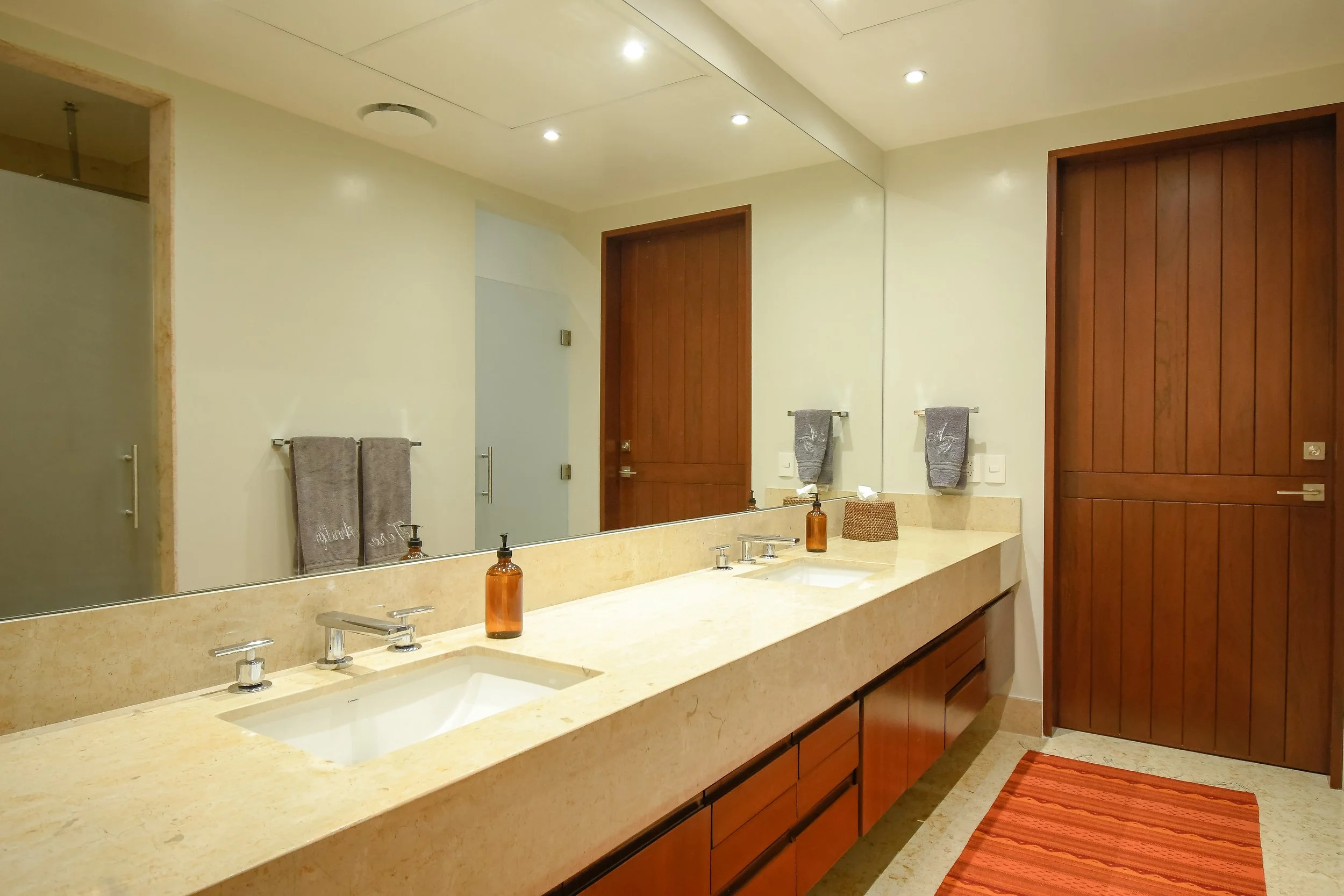 A bathroom with a double sink vanity and large mirror, beige countertop, brown wooden cabinets, gray towels, amber soap dispensers, and a red striped rug.