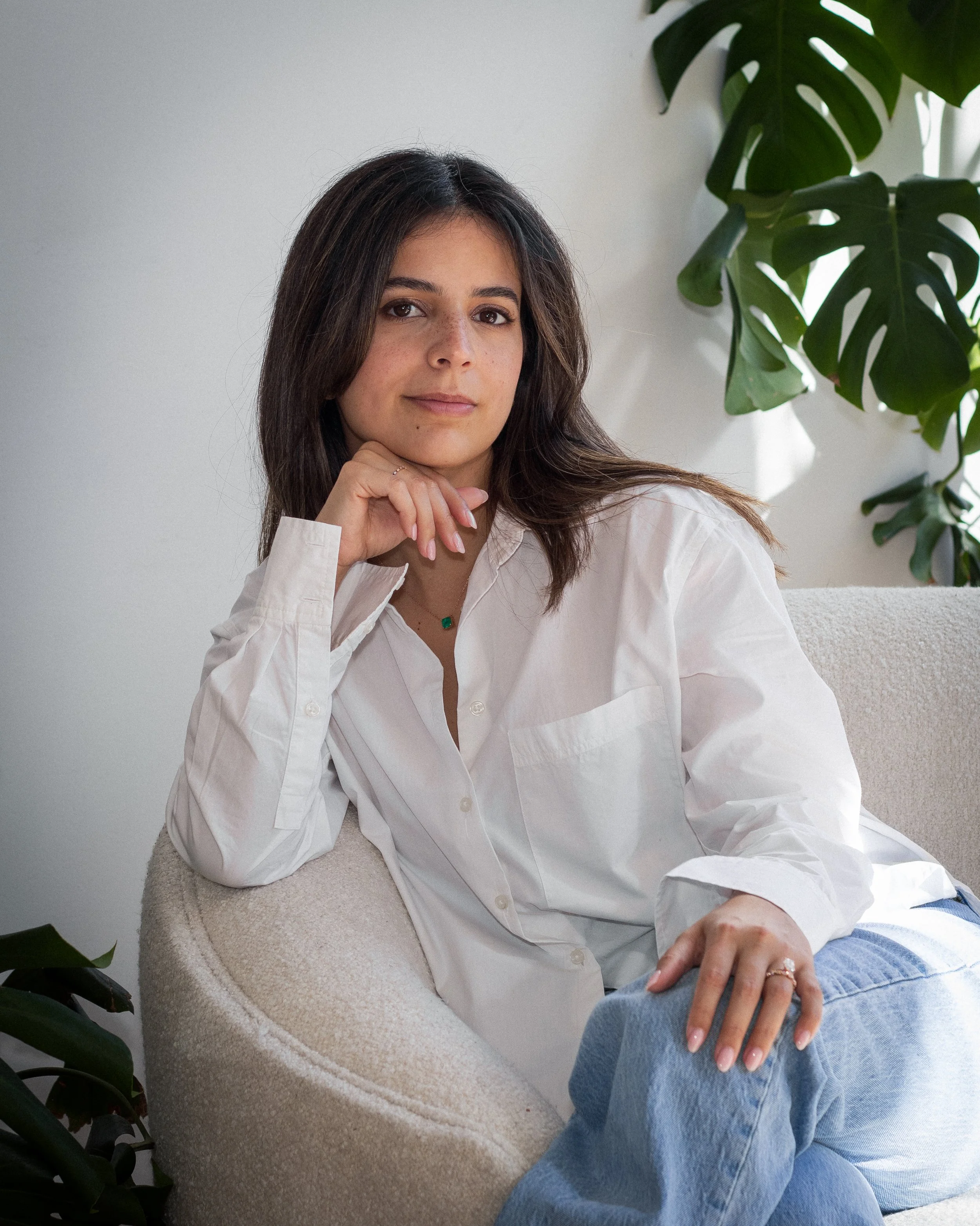 A woman with dark brown hair sitting on a beige textured armchair with a white shirt and light blue jeans, hand resting on her knee, in a room with green houseplants and white walls.