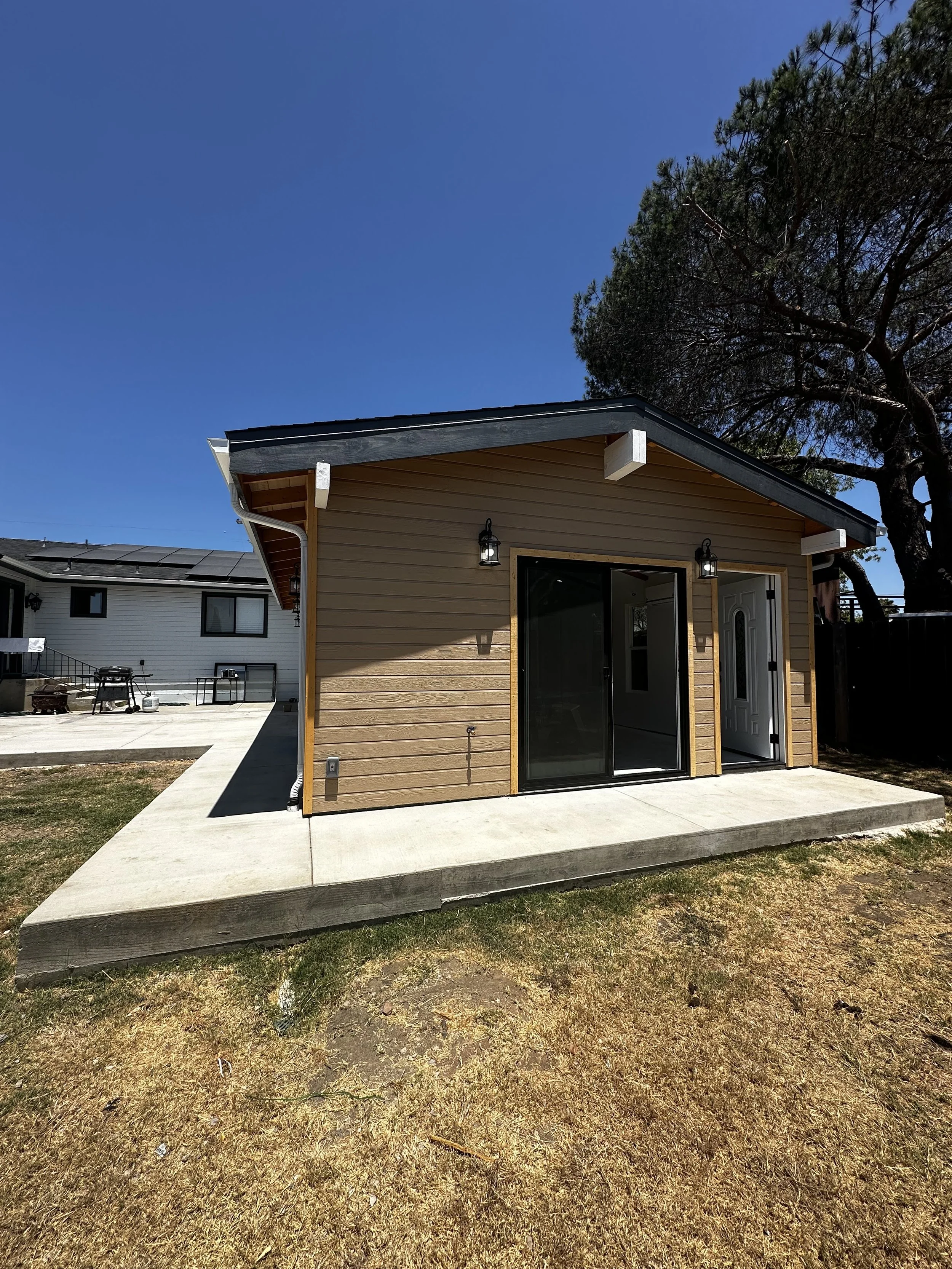 Small backyard building with a sliding glass door and a white door, mounted with two black outdoor lantern lights, on a concrete patio, under a clear blue sky.