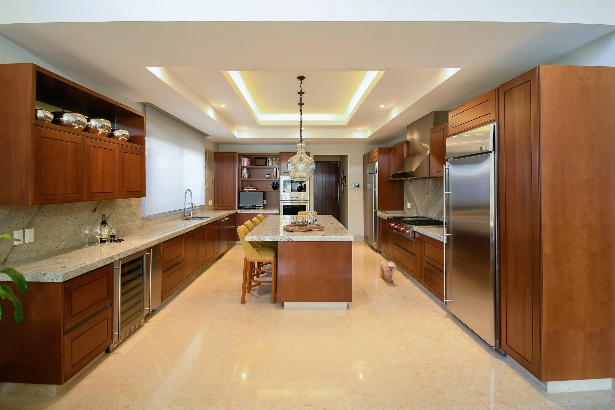 Modern kitchen with wooden cabinets, a large marble-topped island, yellow bar stools, stainless steel appliances, and a small dog on the floor.