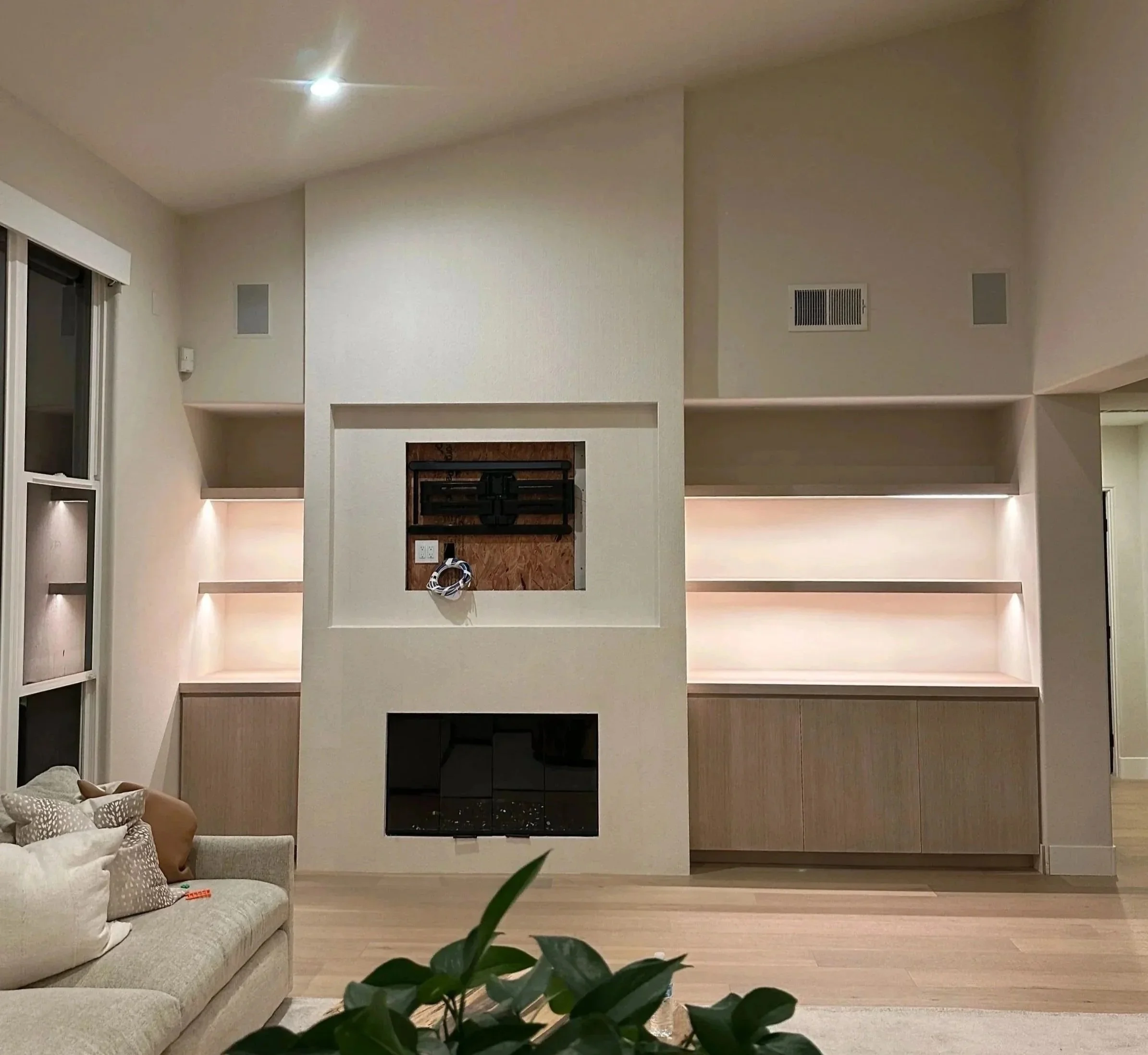 Living room with built-in shelves, a fireplace, and a wall-mounted TV. There are pillows on a tan sofa, and a large window with white curtains on the left. A plant is in the foreground.