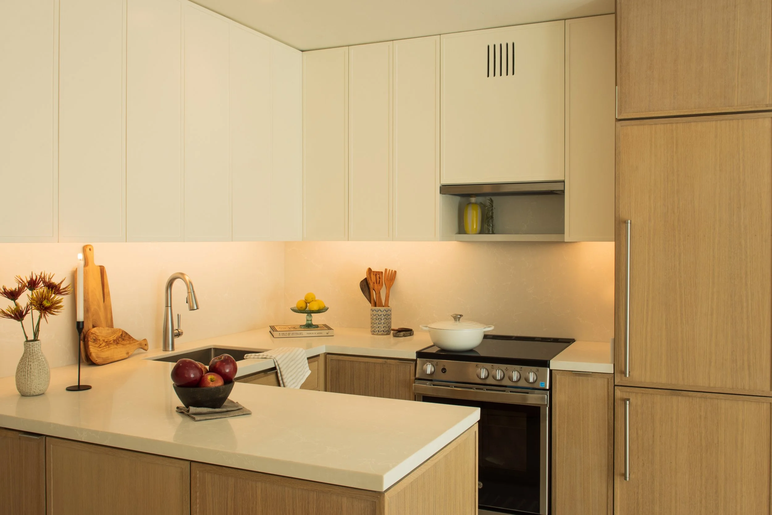 Modern kitchen with light wood cabinets and white countertops, decorated with a vase of flowers, a bowl of apples, and various kitchen utensils, with a stove and oven.
