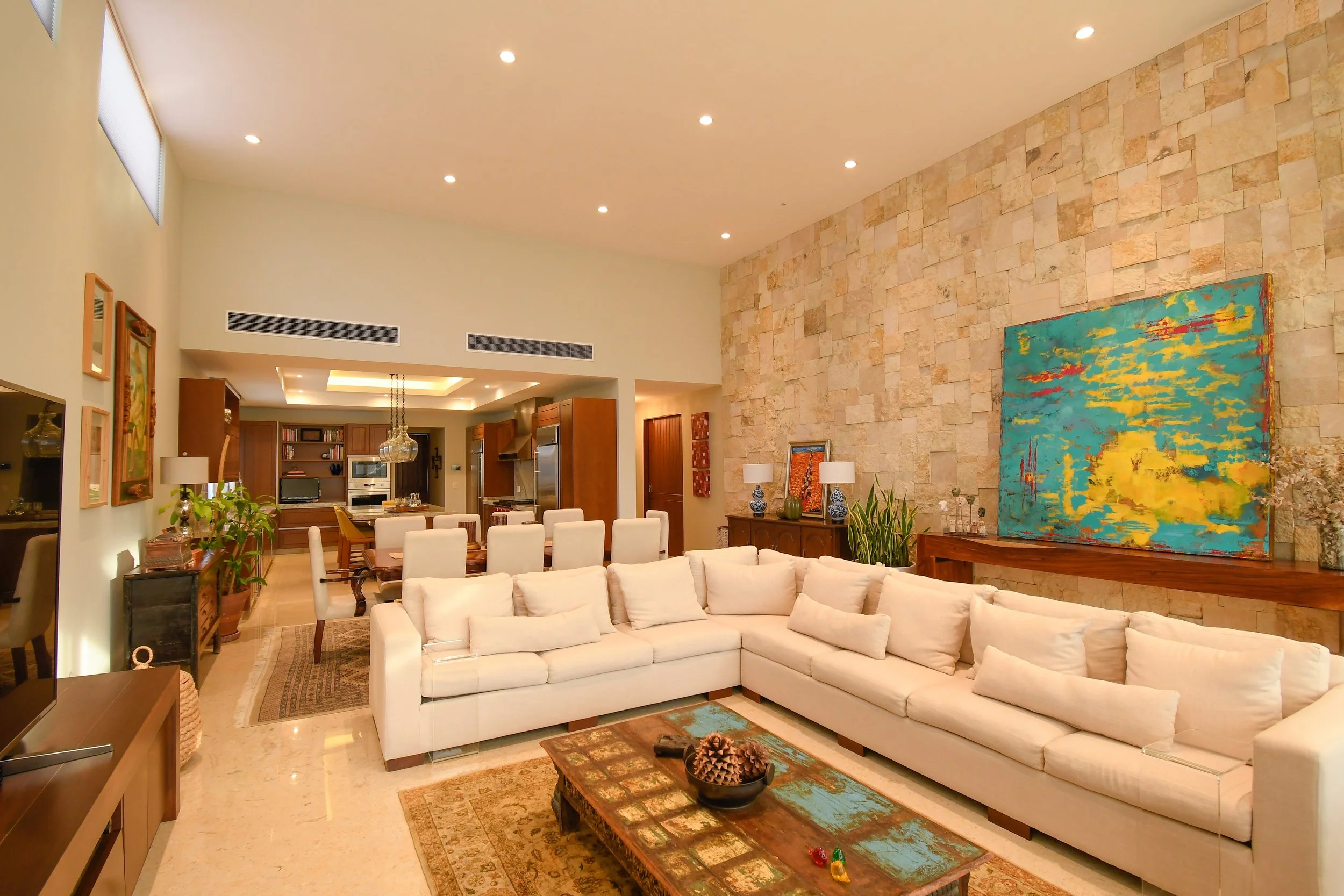 Spacious living room with white sectional sofa, stone accent wall, colorful abstract painting, wooden coffee table with pinecones, and a nearby dining area and kitchen.