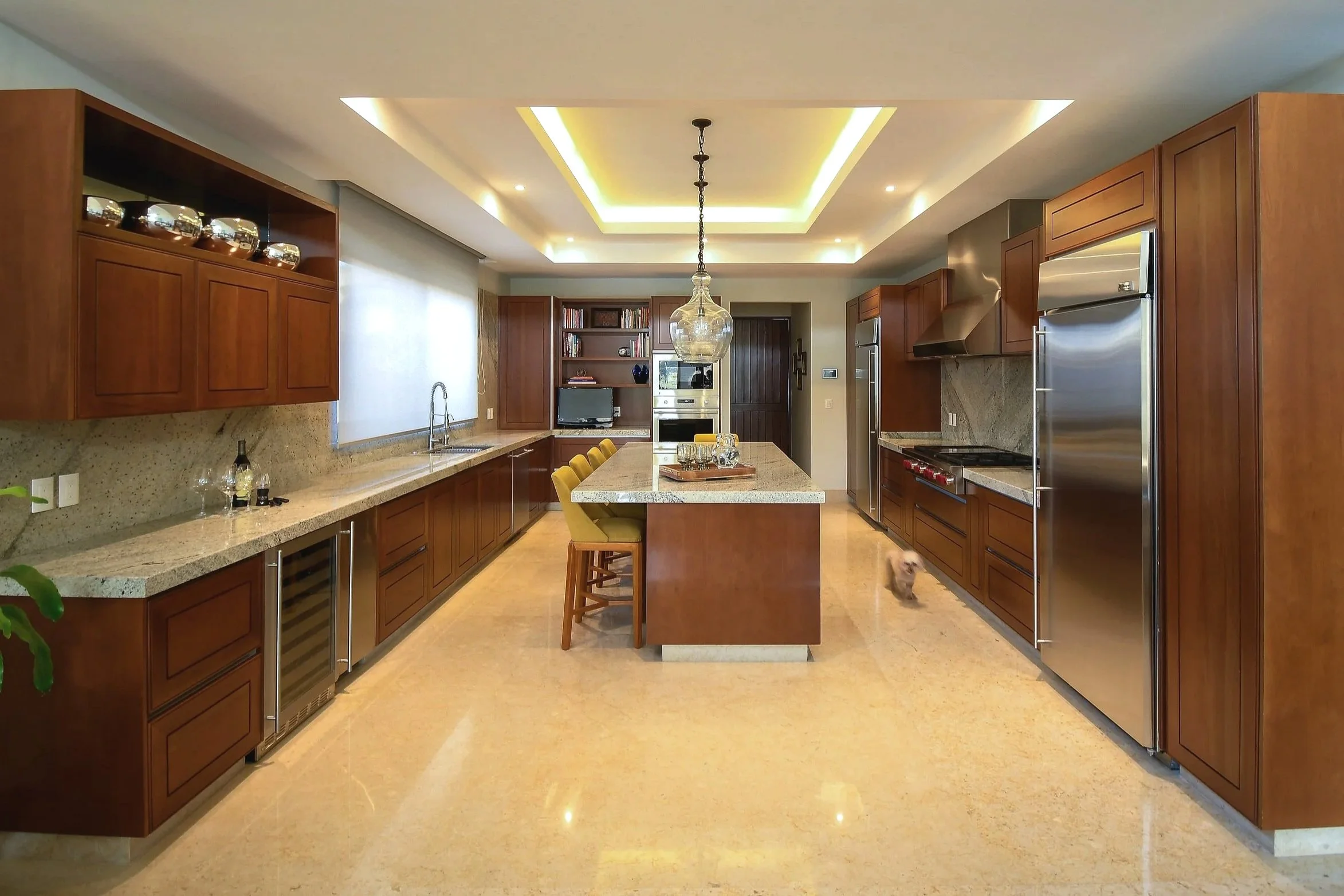 Spacious kitchen with wooden cabinets, granite countertops, stainless steel appliances, and a large central island with seating, illuminated by a chandelier and recessed lighting, with a dog near the right side.