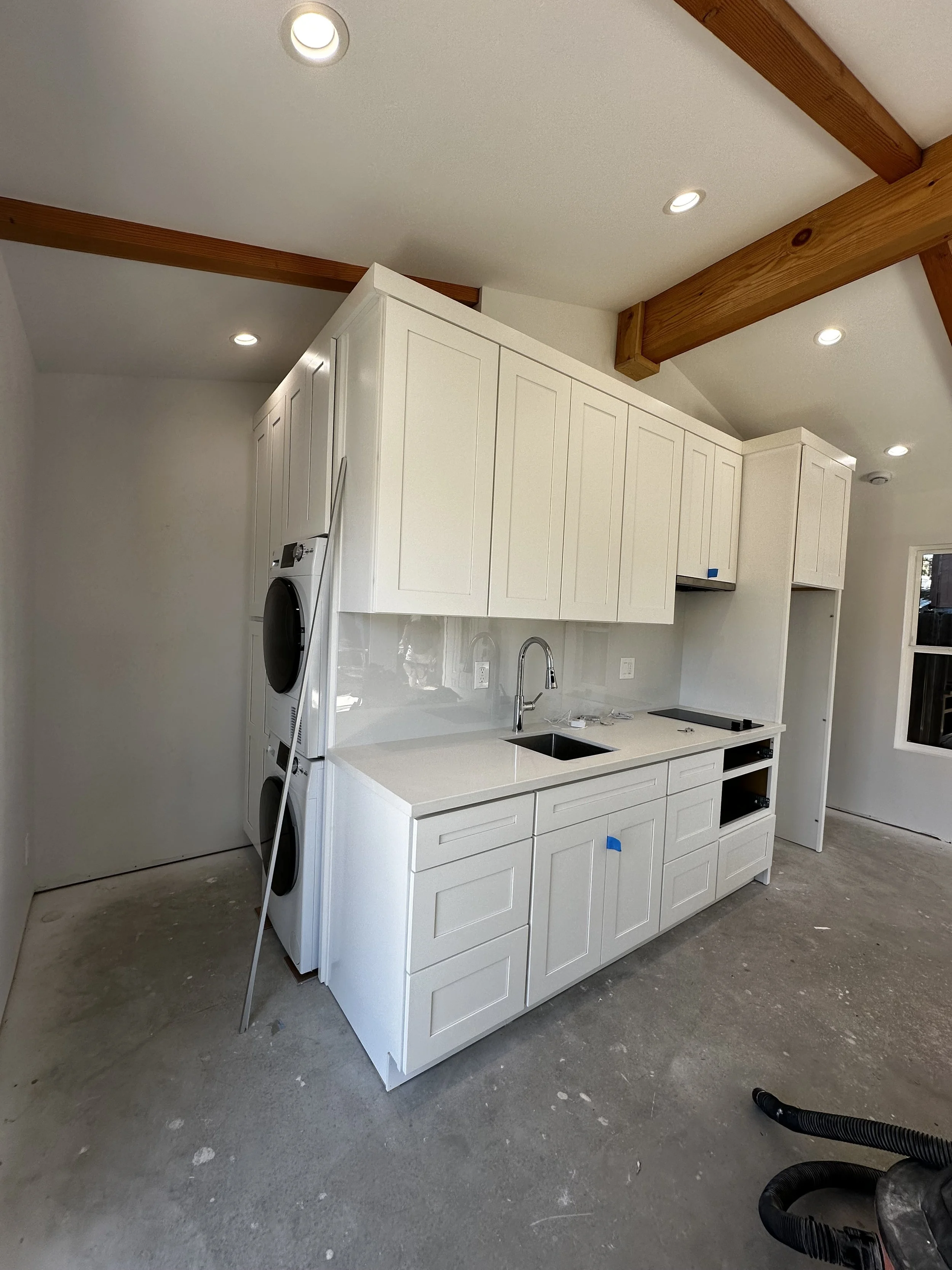 Kitchen under construction with white cabinets, a small sink, and a laundry area with stacked washer and dryer. The floor is unfinished concrete, and there are wooden ceiling beams and recessed lighting.