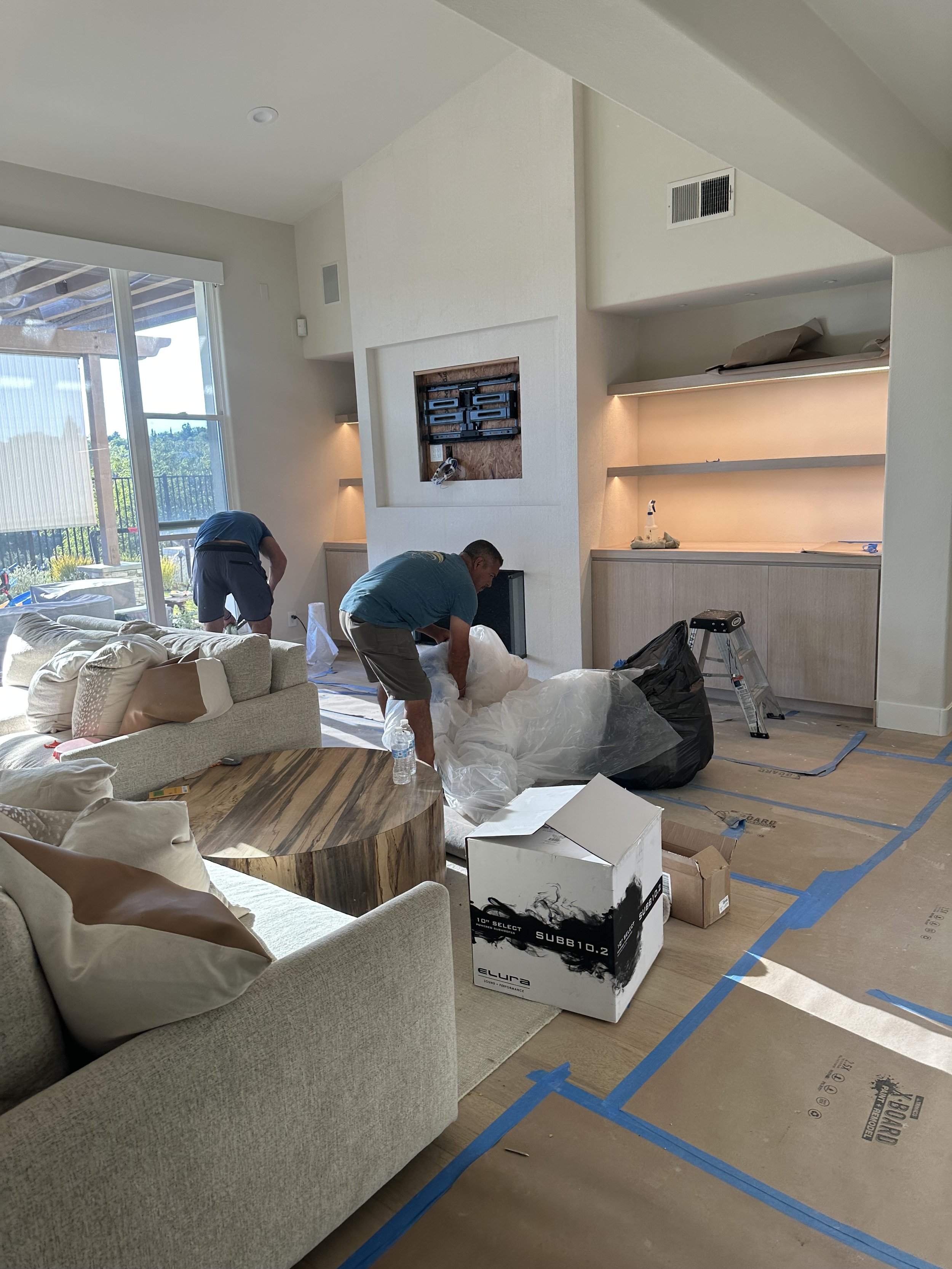 People working in a living room during home renovation, with furniture, packaging, and construction materials visible.