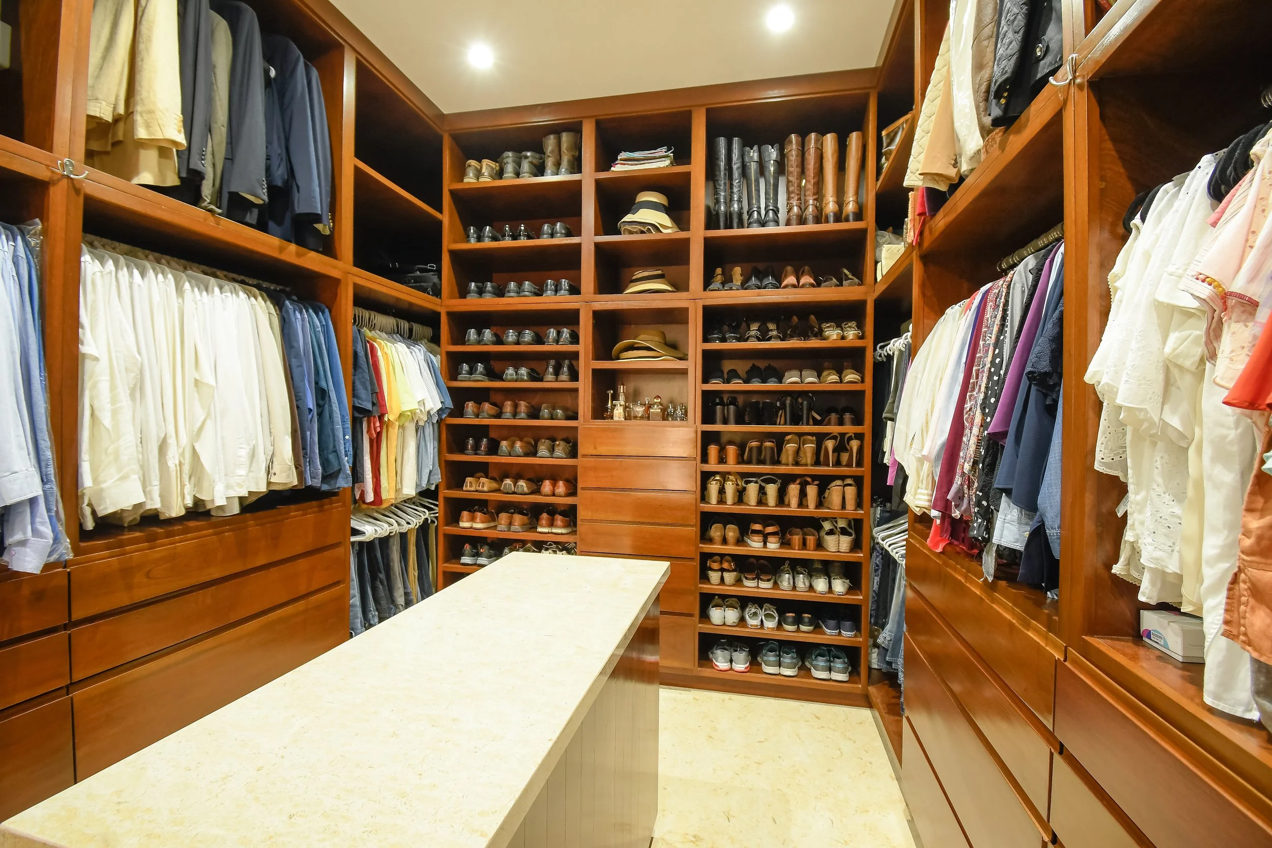 A walk-in closet with wooden shelves filled with shoes, hats, and folded clothing. Clothes hang on both sides of the room, and a large central counter is in the middle.